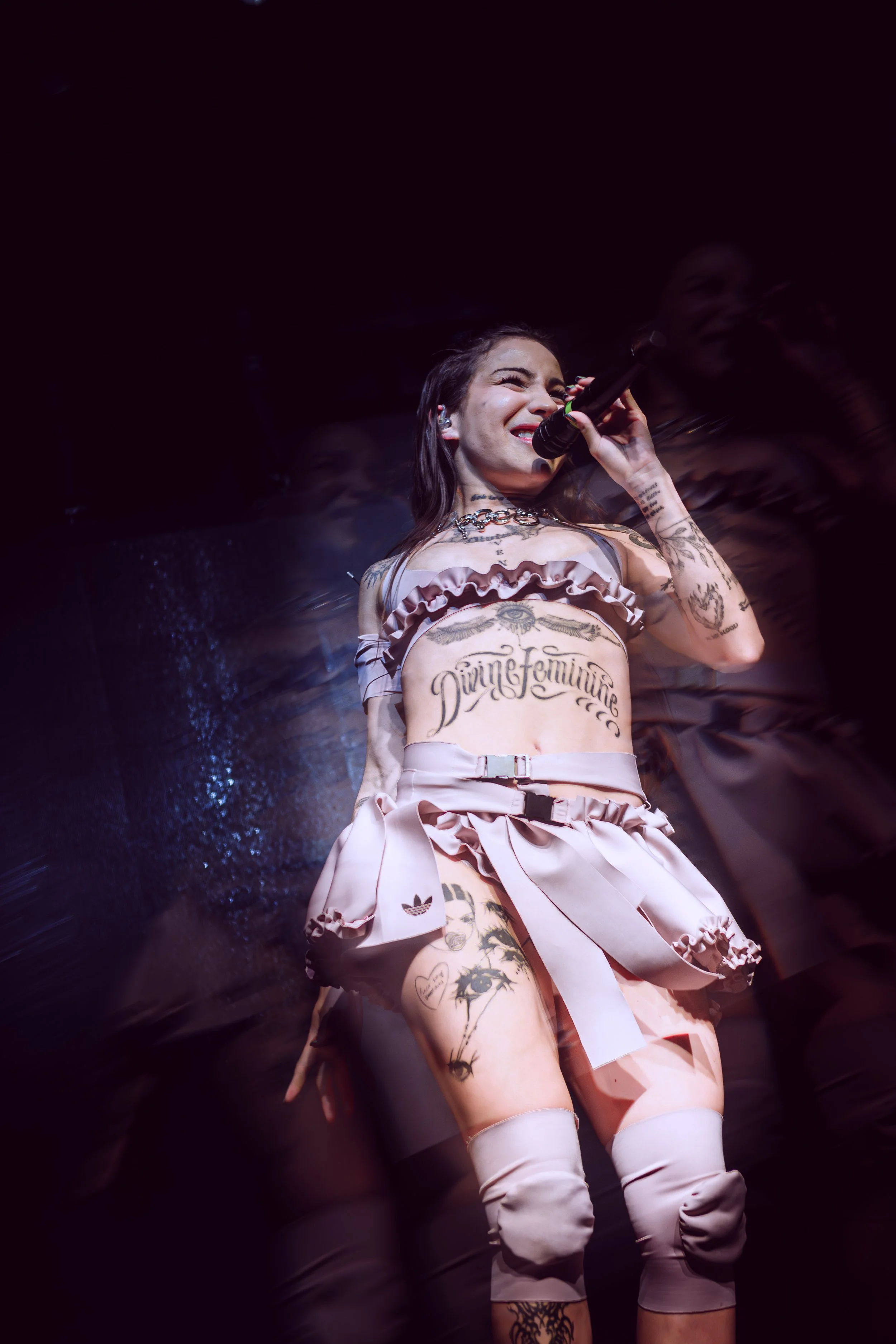 Una mujer cantando en el escenario con tatuajes visibles, vestimenta color rosa y fondo oscuro.
