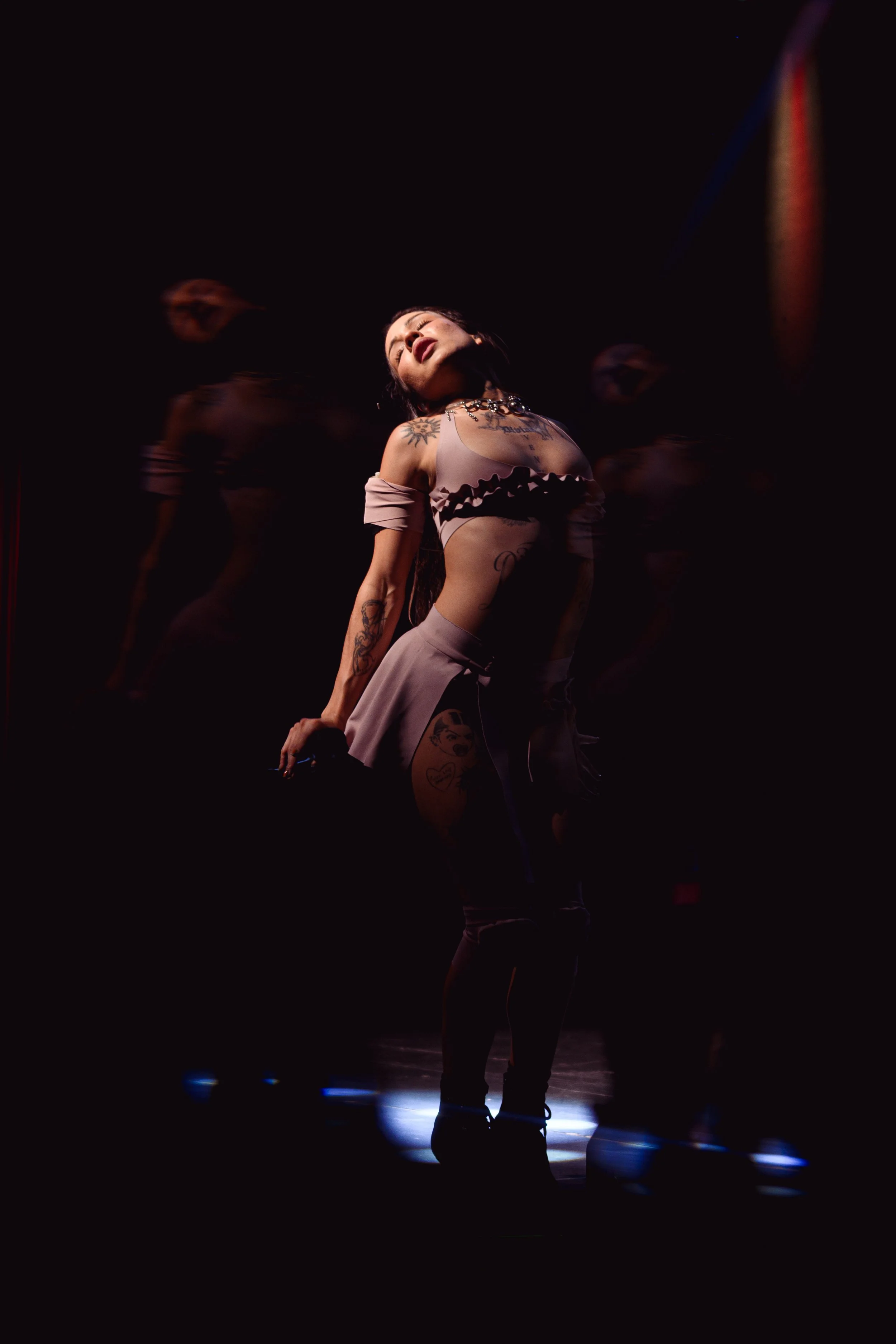 Una mujer con tatuajes en un escenario oscuro, en una pose expresiva con la cabeza inclinada hacia atrás y los ojos cerrados, iluminada por una luz focal.