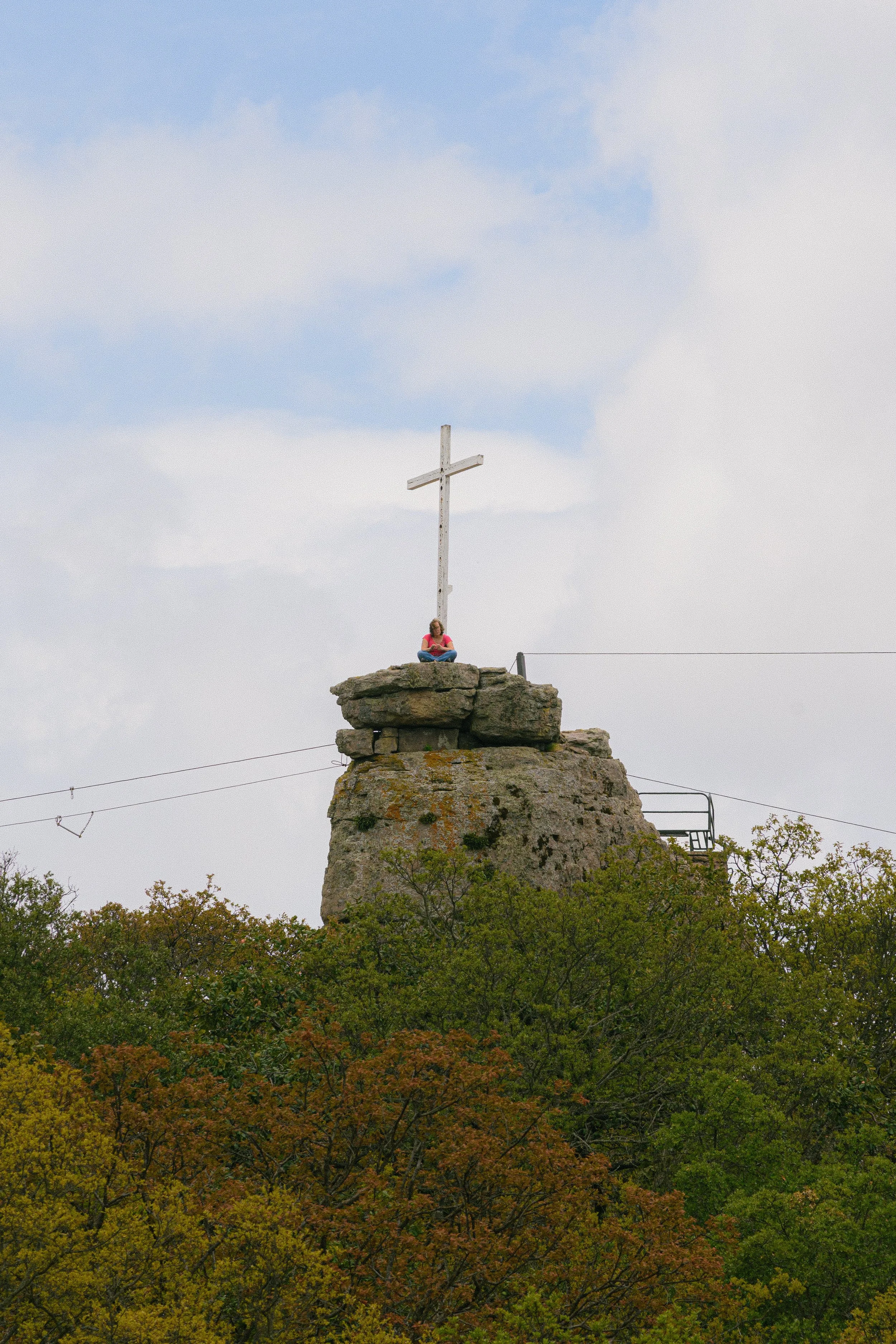 Una mujer sentada en una roca en la cima de una roca grande, con una cruz de madera detrás de ella y árboles frondosos en primer plano.