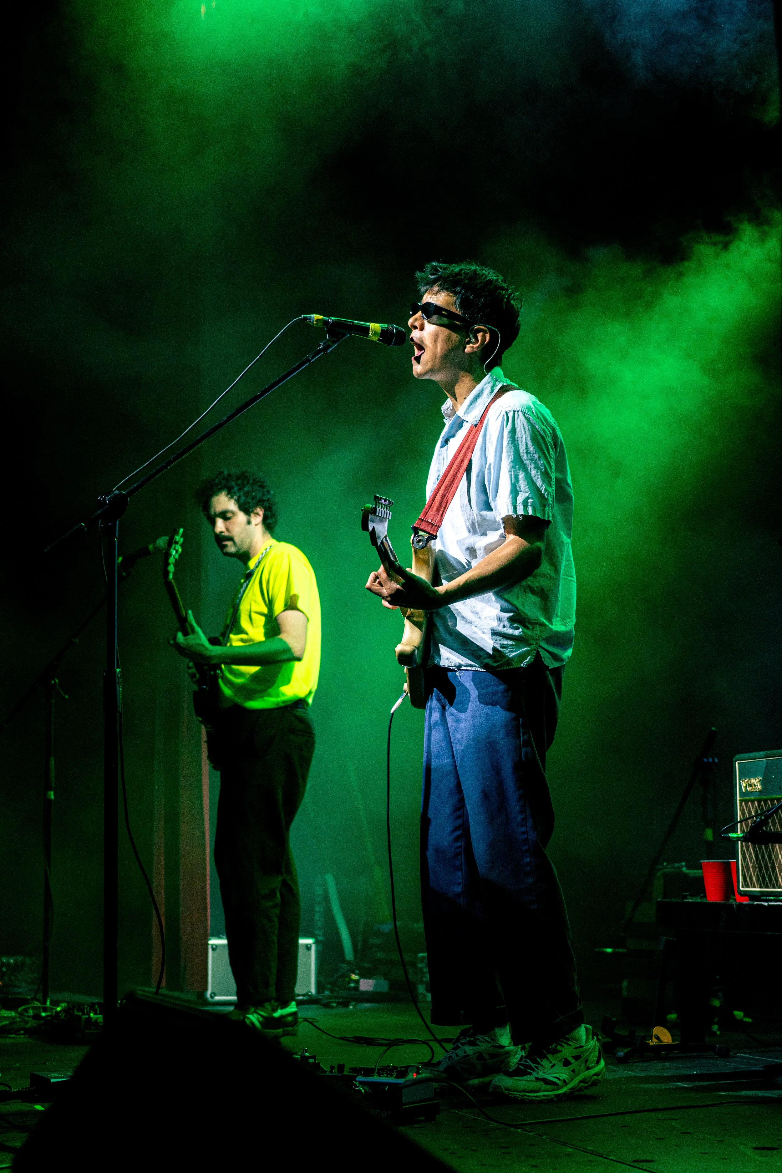 Dos músicos tocando en un escenario con iluminación verde, uno con gafas oscuras y guitarra eléctrica, el otro con guitarra y camiseta amarilla.