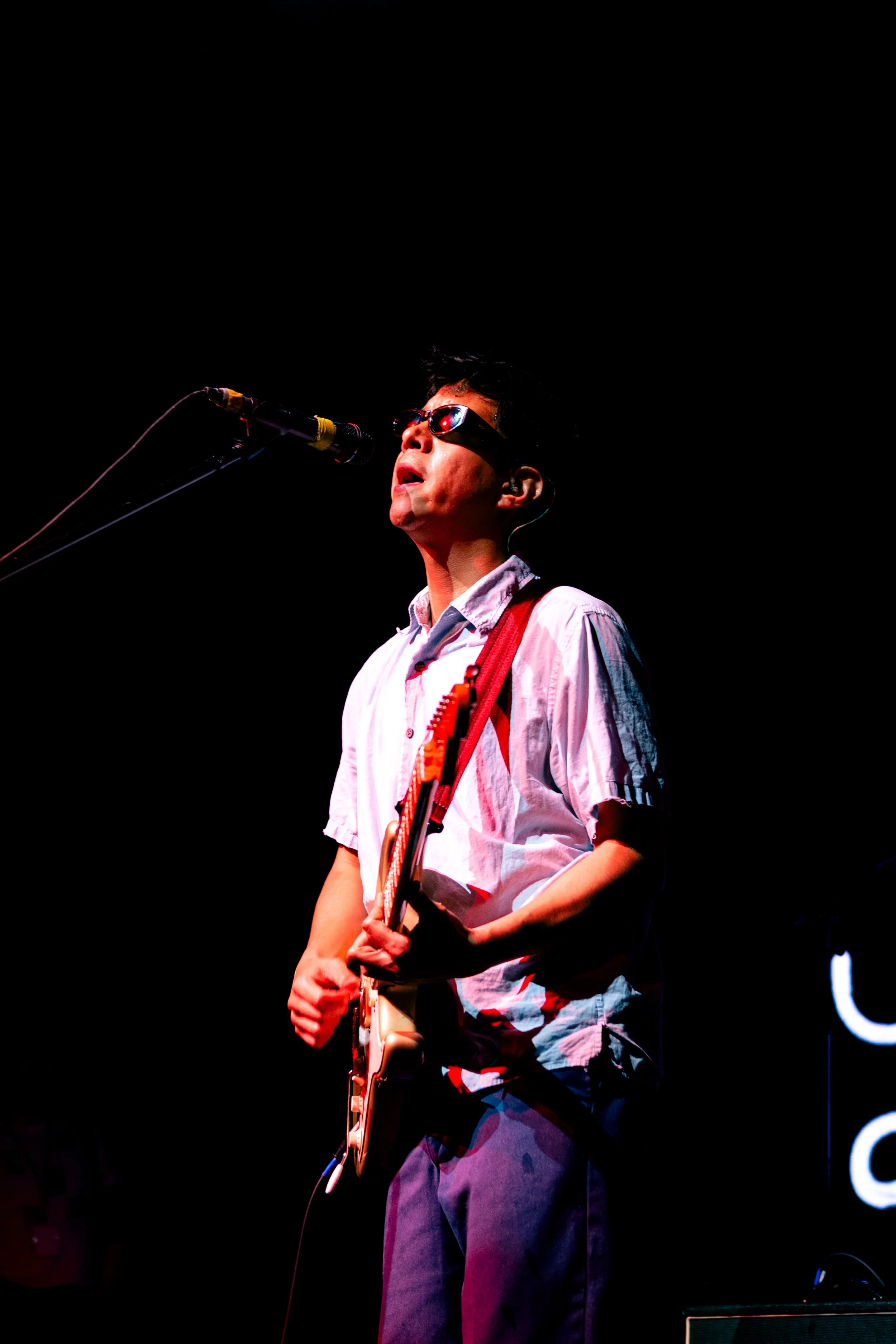 Joven tocando guitarra eléctrica y cantando en un concierto, usando gafas de sol y camisa blanca, con fondo negro.