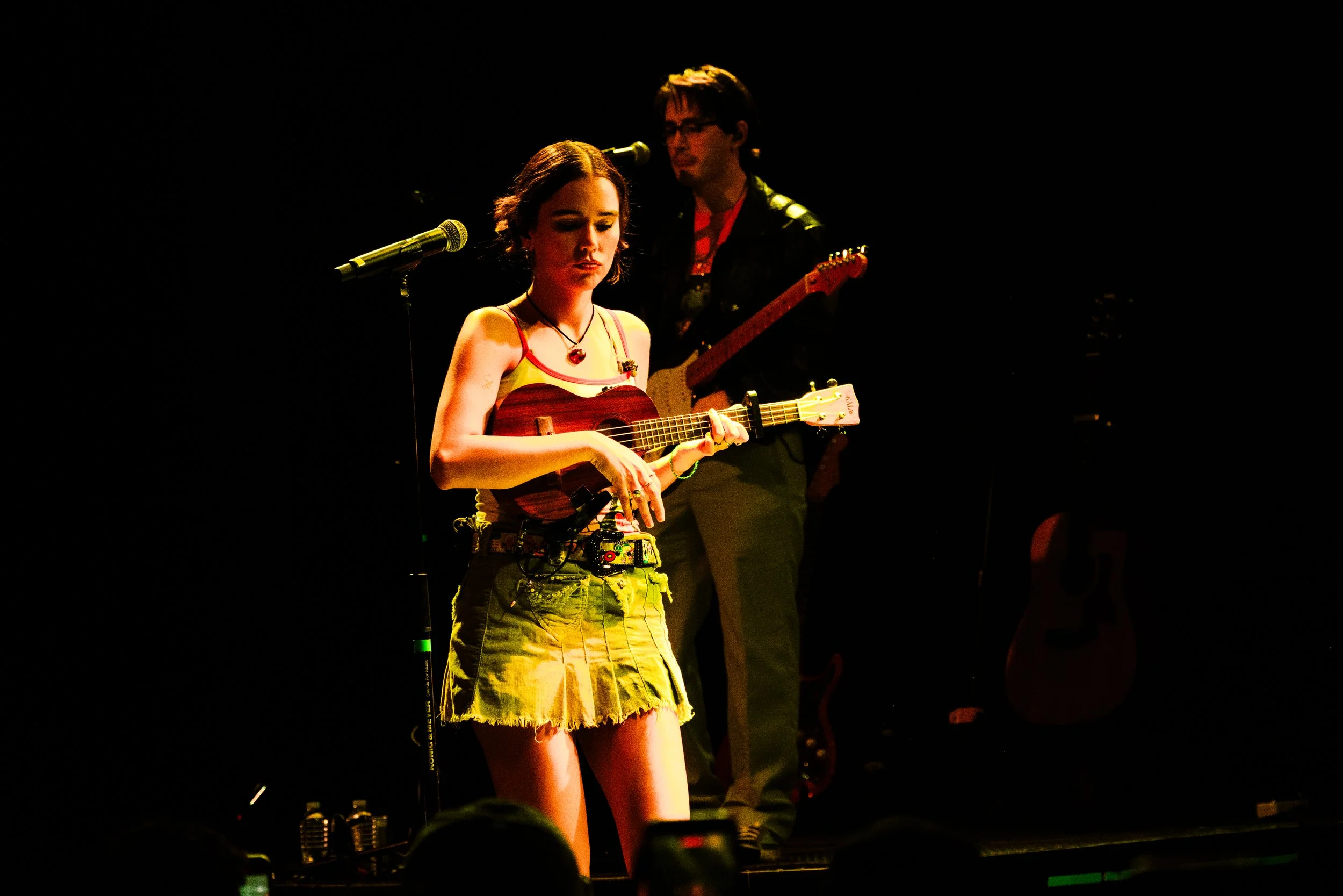 Una mujer tocando la guitarra en un escenario con un hombre al fondo tambien tocando la guitarra.
