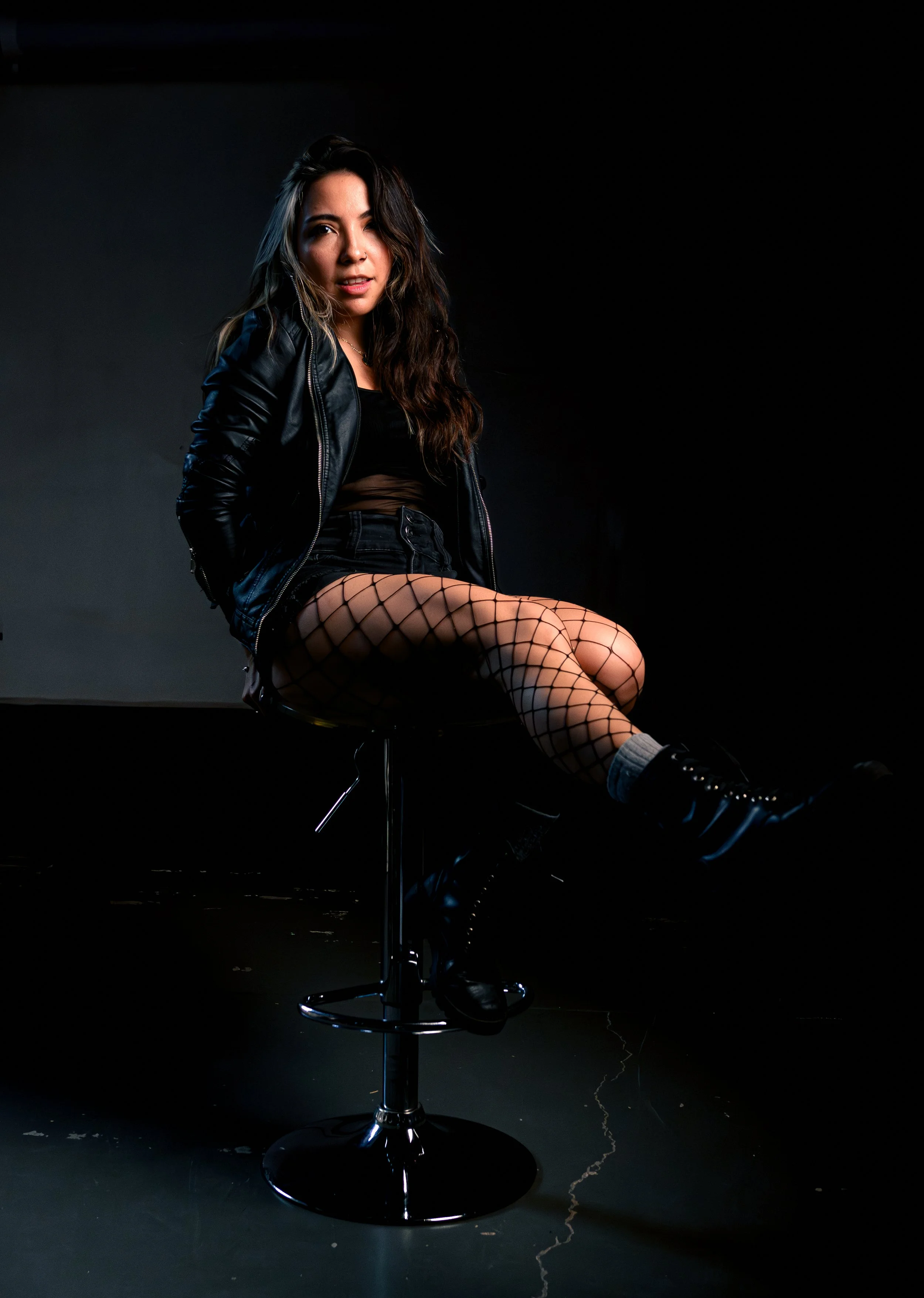 Joven mujer con cabello rizado sentada en una silla de escritorio, vistiendo una chaqueta de cuero, falda corta y medias de red, en un fondo negro.