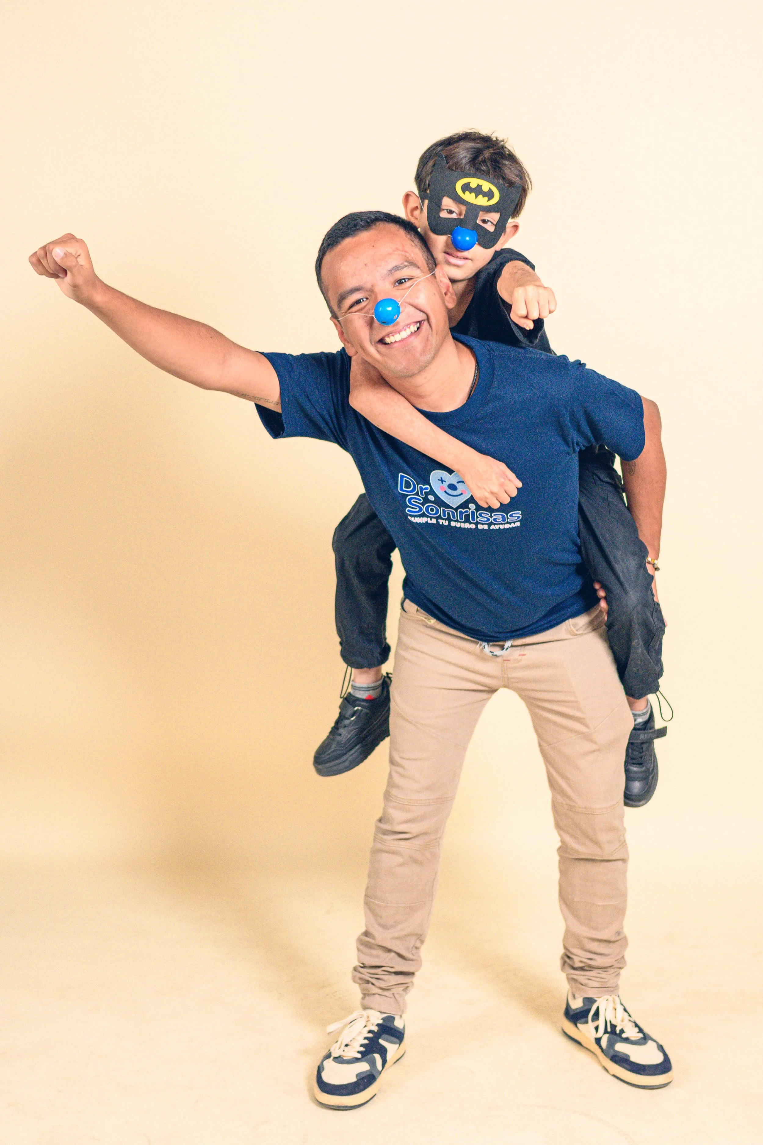 Un hombre y un niño con narices y máscaras de payaso y Batman, en un fondo beige, mostrando alegría y diversión.