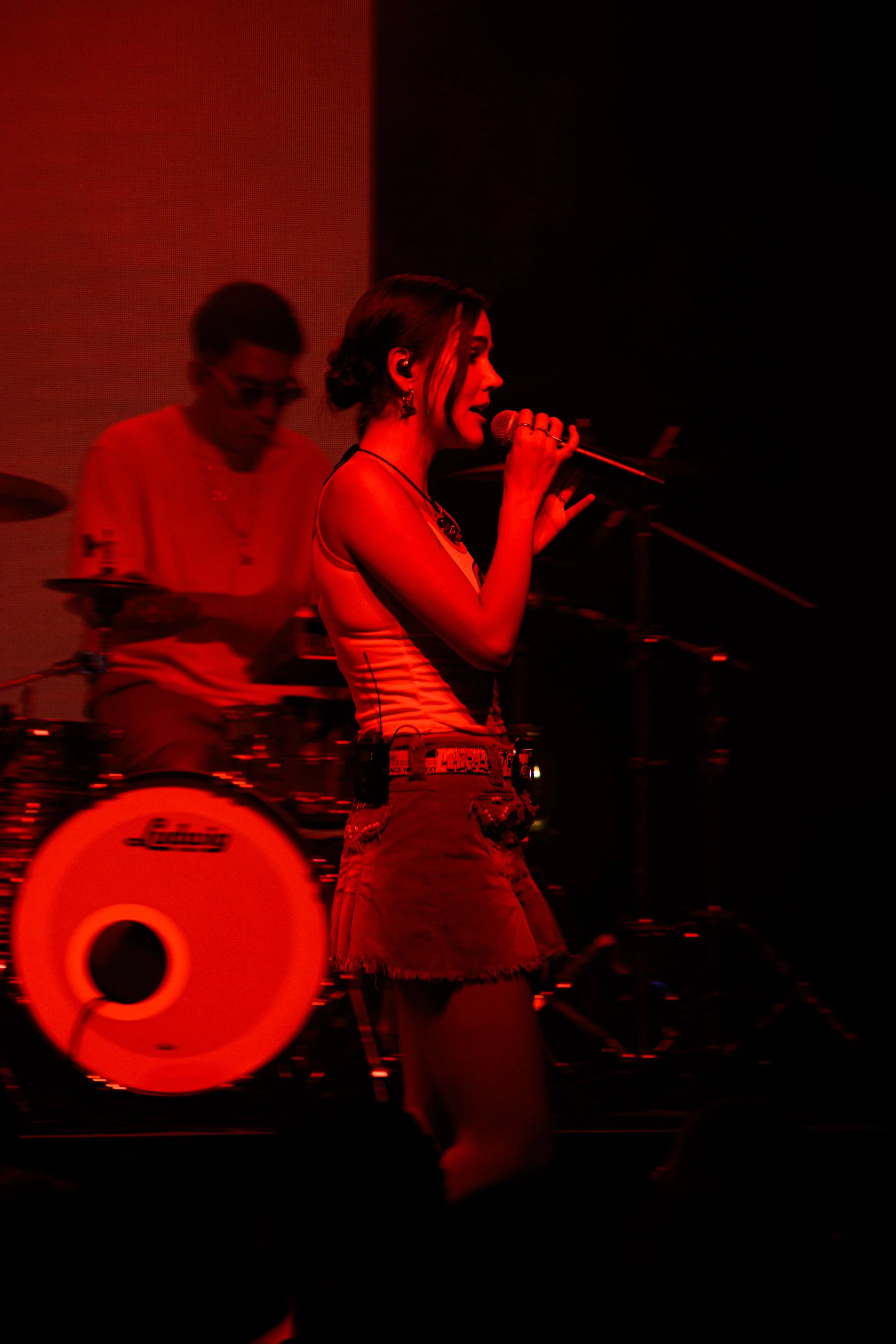 Joven mujer cantando en un concierto, rodeada de luces rojas, con un músico tocando la batería en segundo plano.