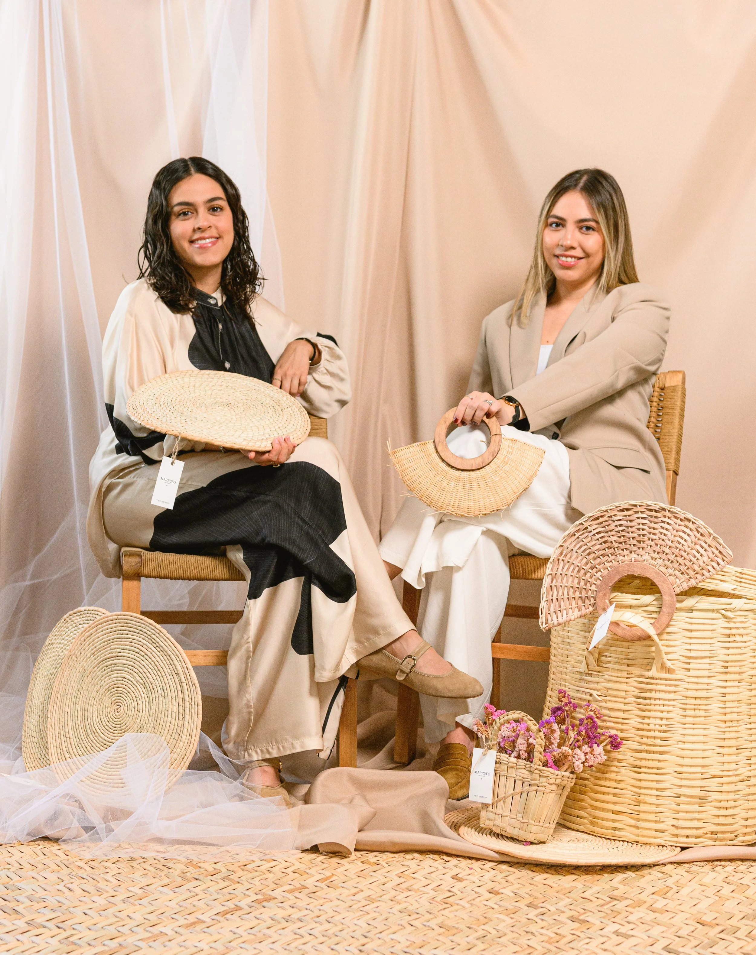 Dos mujeres sentadas en sillas de mimbre, rodeadas de sombreros de paja y arreglos florales, en un entorno decorativo con cortinas beige.