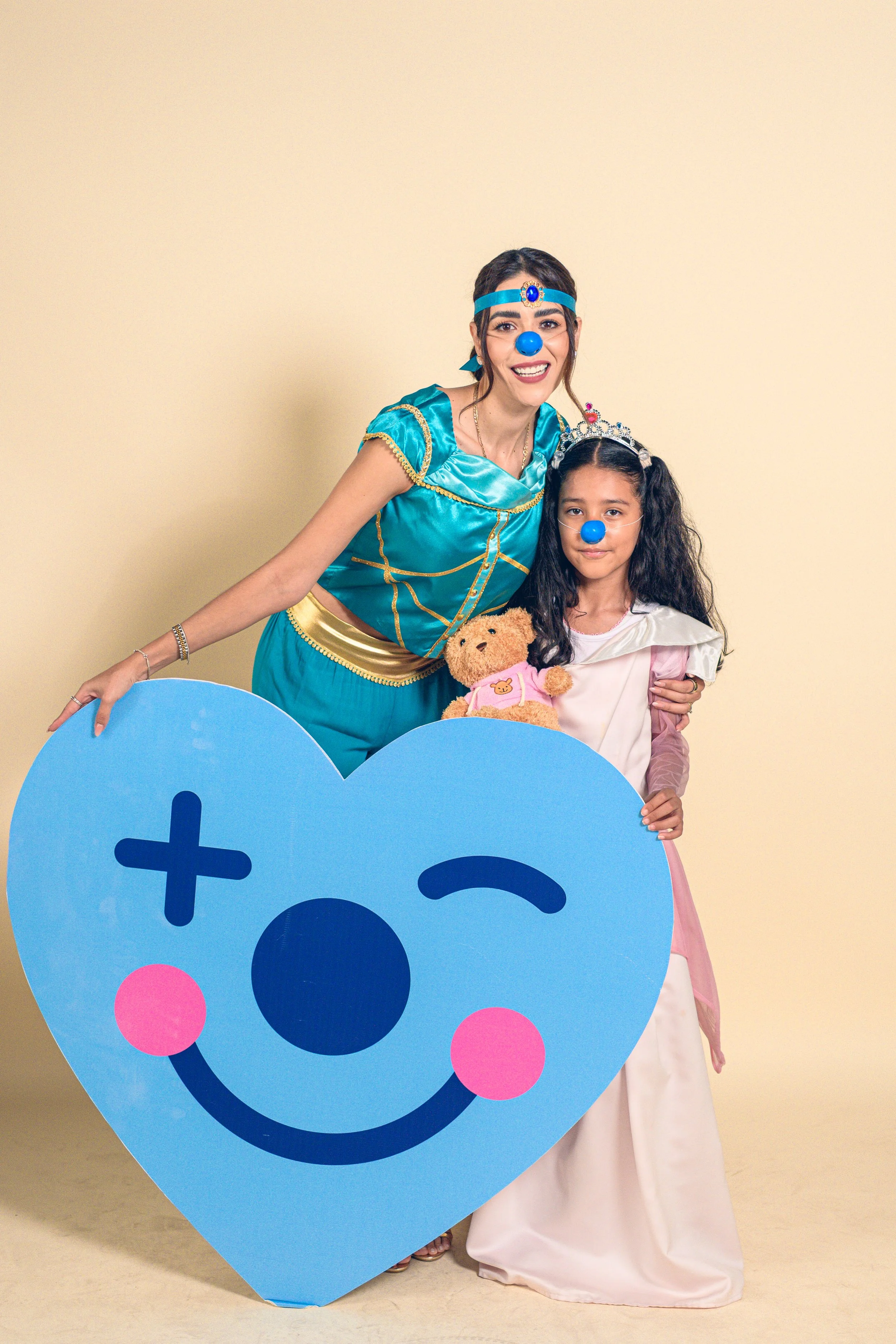 Una mujer y una niña disfrazadas, ambas con narices y caritas de payaso, sosteniendo un corazón grande con cara de emoticón en tonos azules y rosados, en un fondo beige.