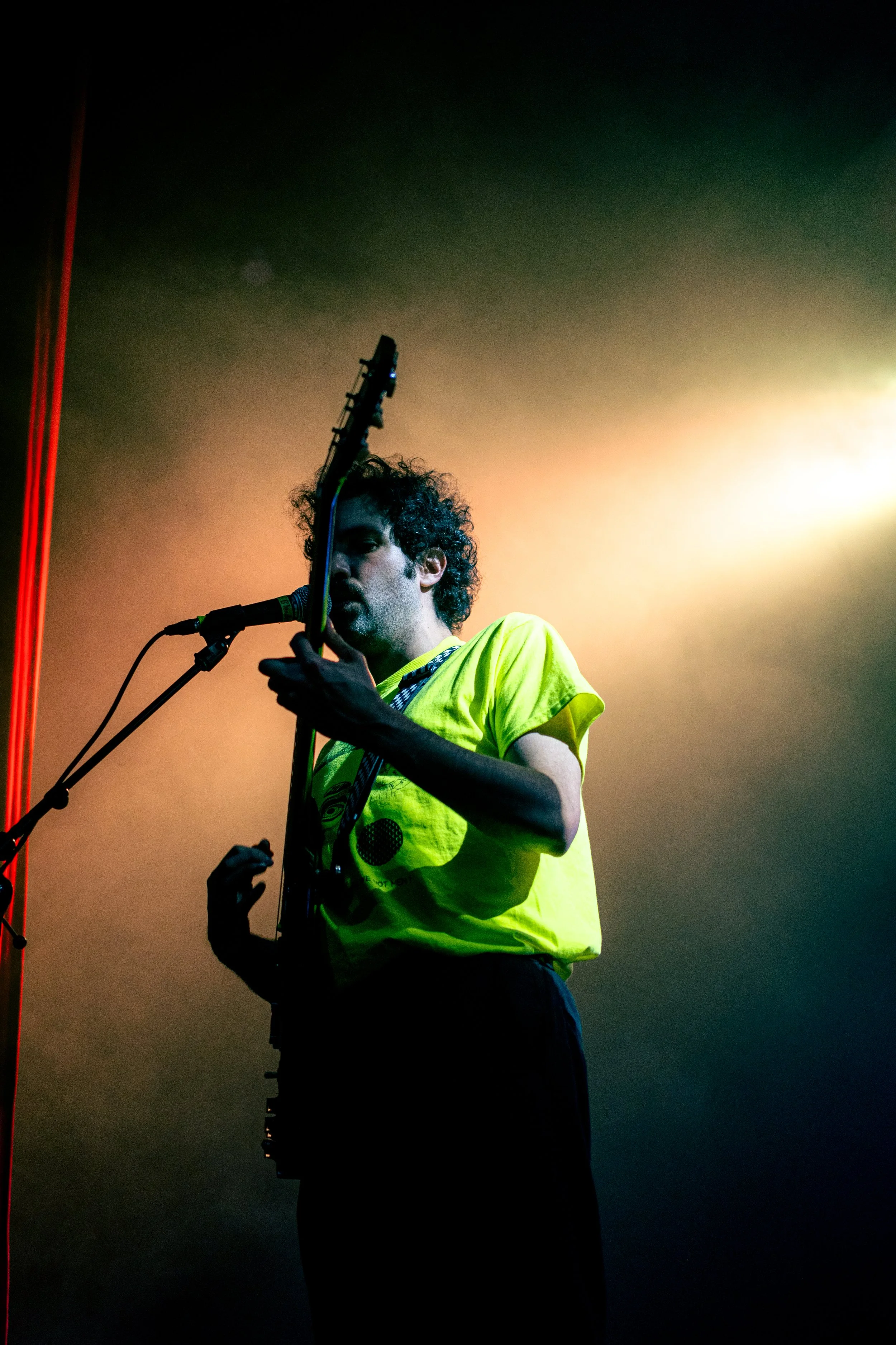 Hombre tocando guitarra y cantando frente a micrófono, con fondo difuso de luces de escenario.