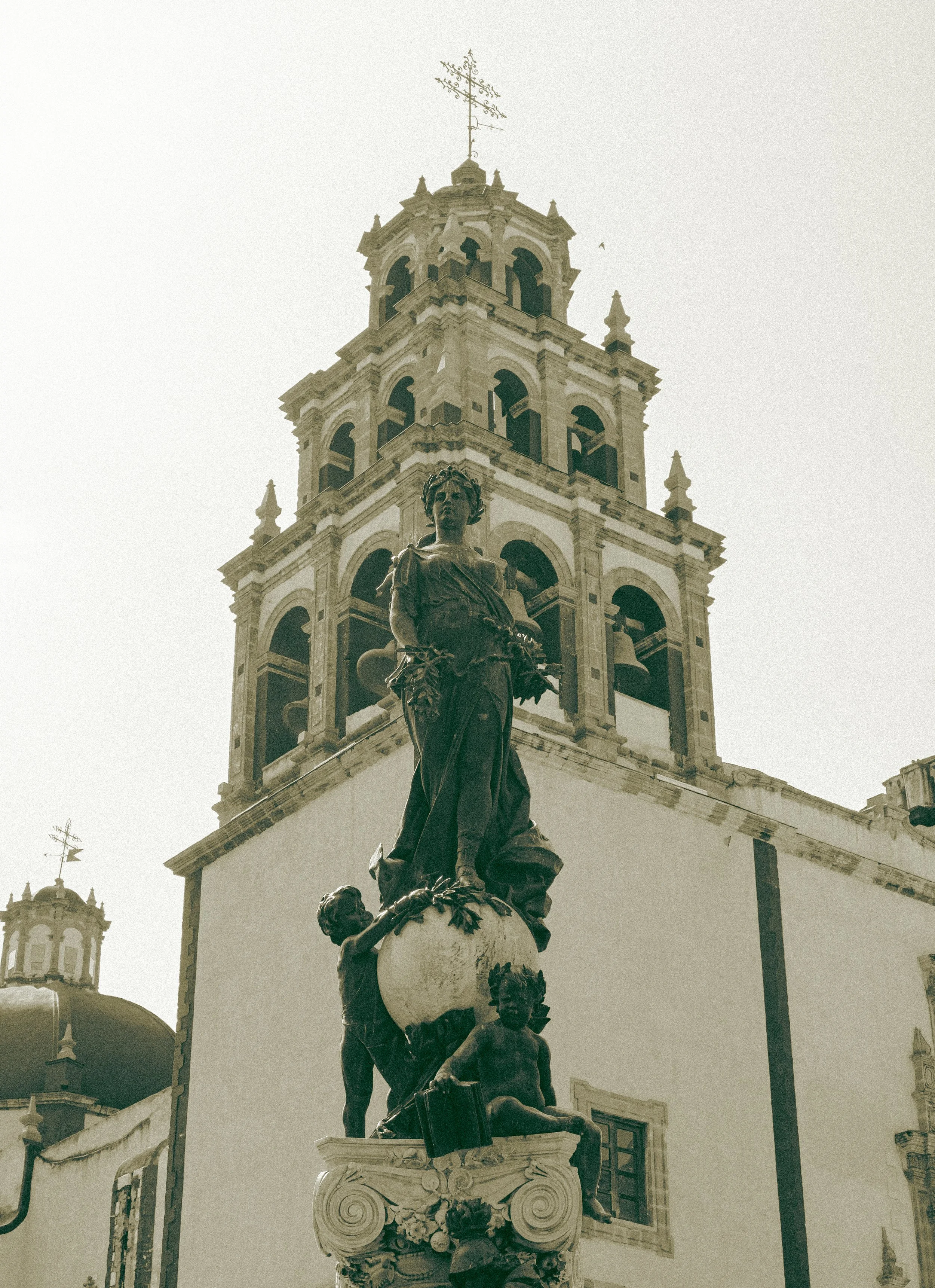 Escultura en un pedestal frente a un edificio con campanario, probablemente en una ciudad colonial.