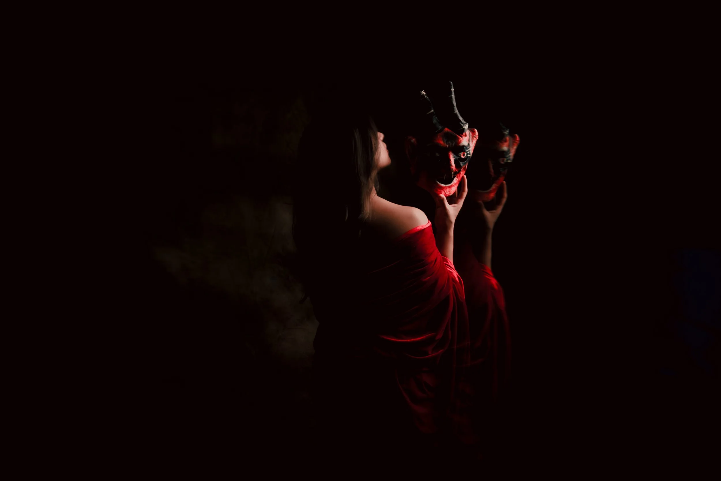 Una mujer con vestido rojo sostiene dos máscaras de diablo en un fondo oscuro.