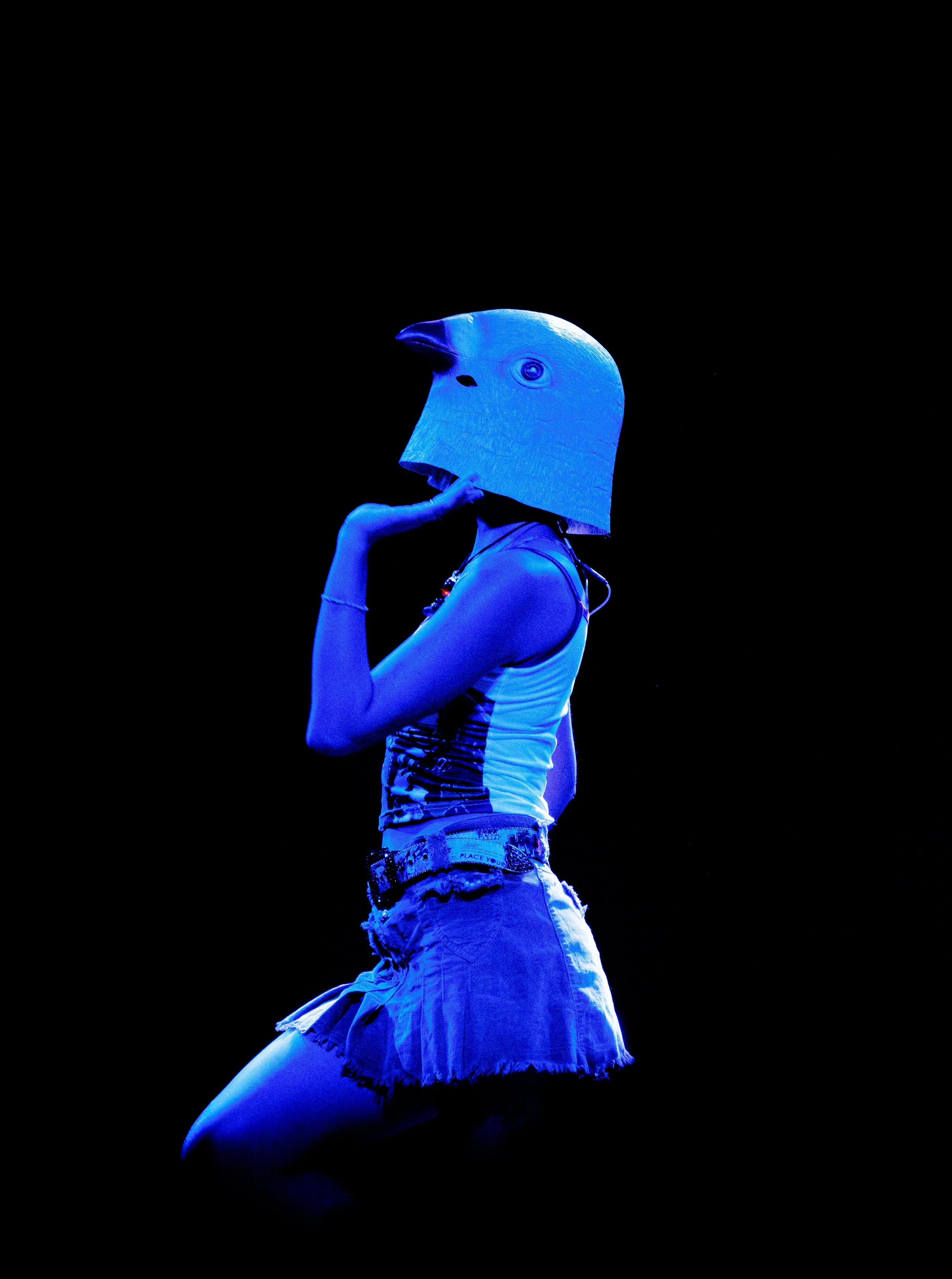 Persona con falda y camiseta, usando un casco con forma de cabeza de ave en un fondo negro.