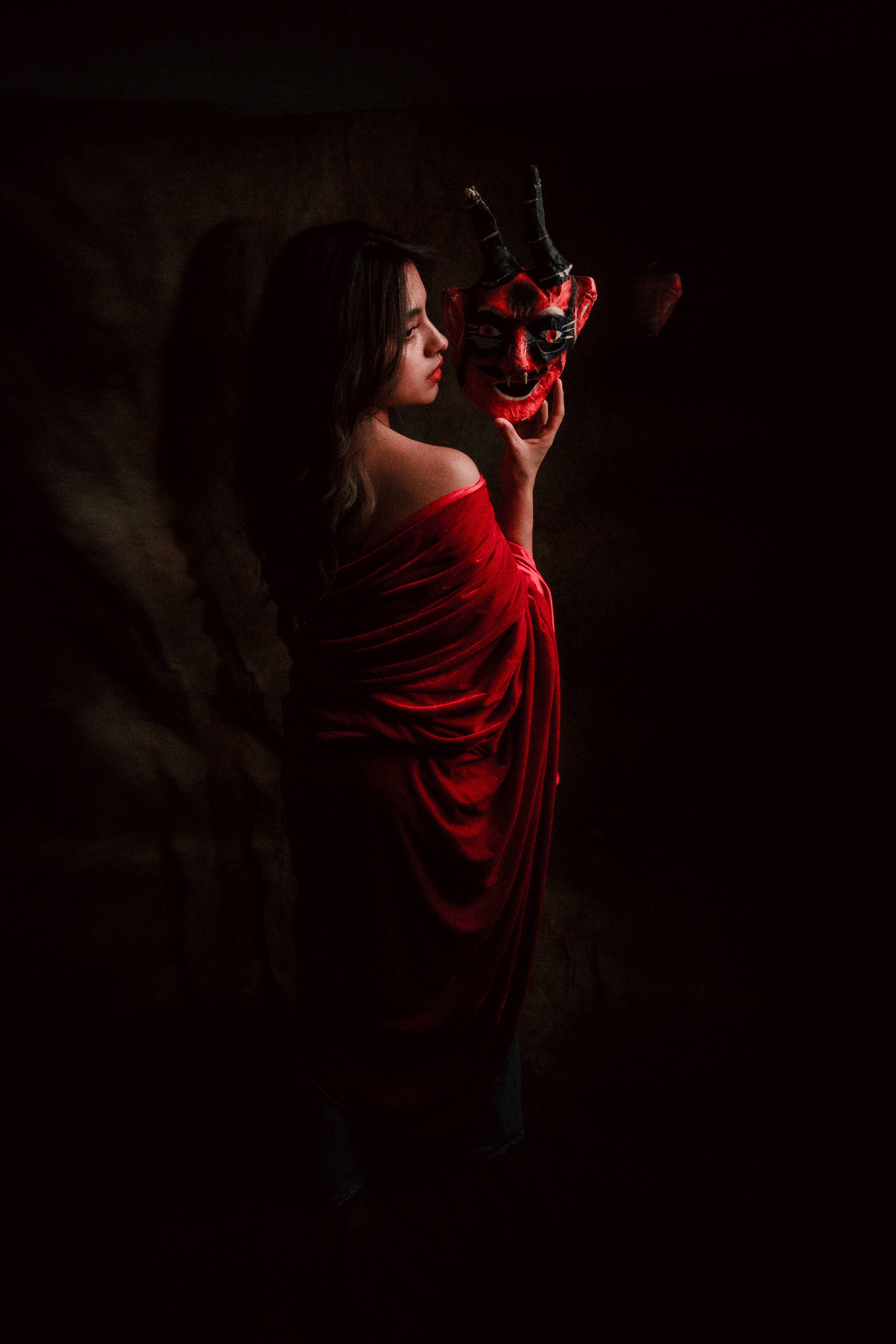 Mujer con vestido rojo sostiene una máscara de diablo roja y negra en un fondo oscuro.