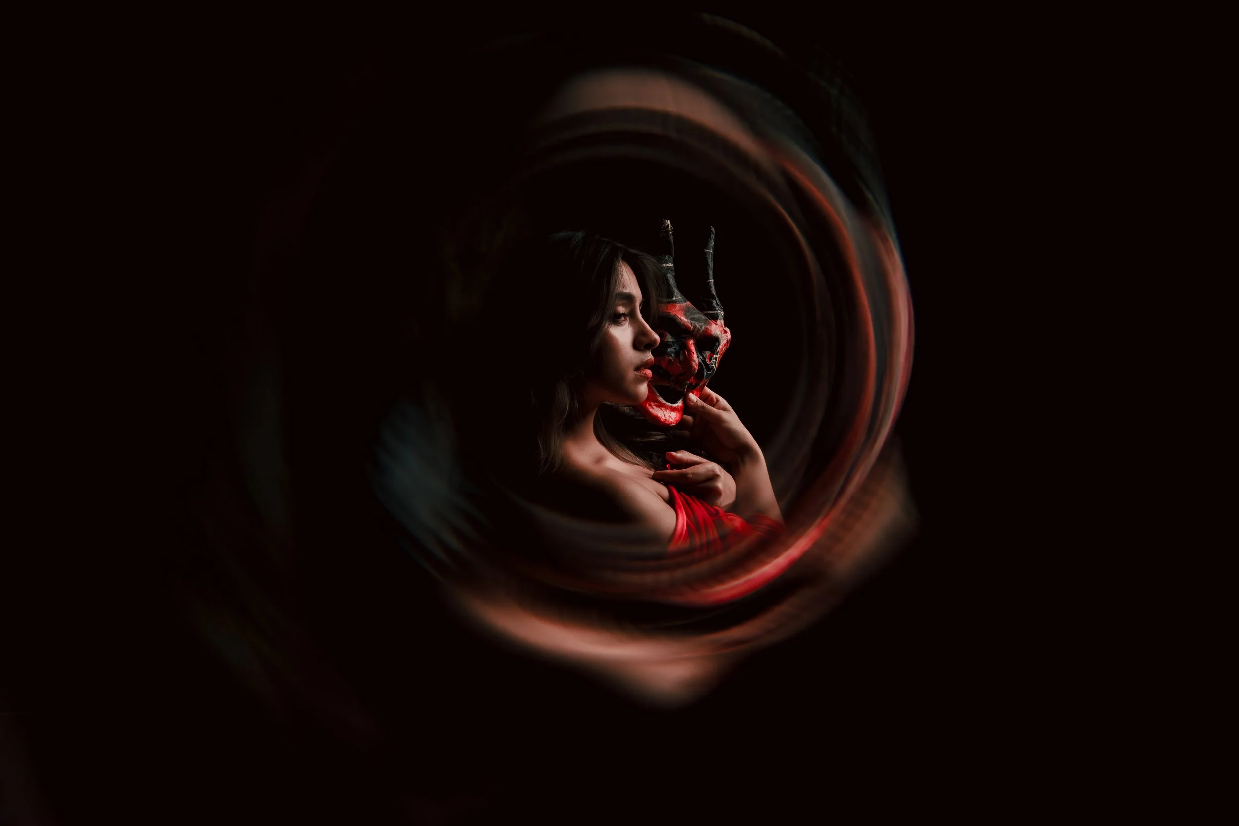 Mujer con cabello largo y vestido rojo sosteniendo una máscara de demonio con cuernos en un entorno oscuro y misterioso.