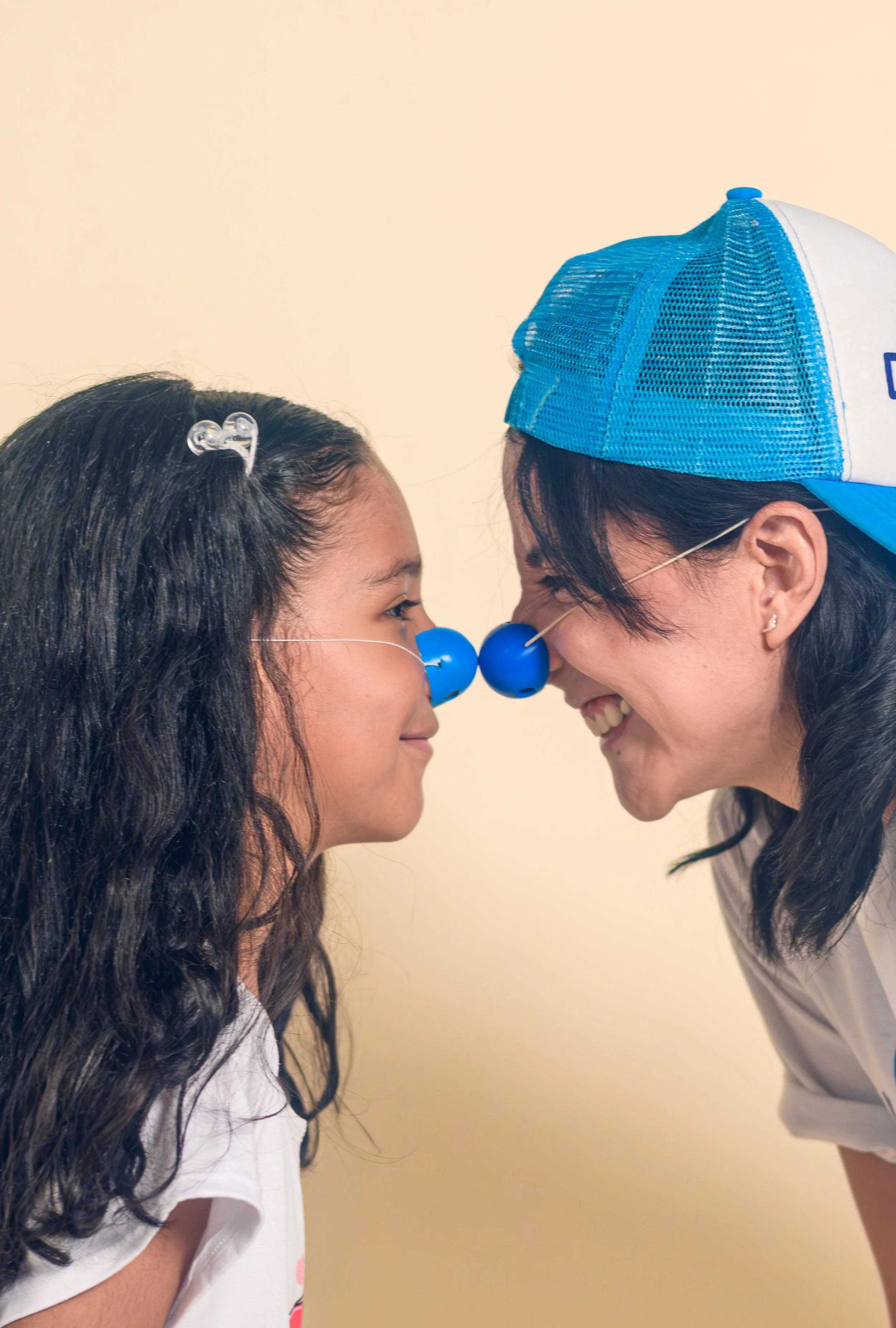Una niña y una mujer con narices de payaso, tocándose la punta de la nariz, sonriendo y con la frente tocándose, en un fondo beige.