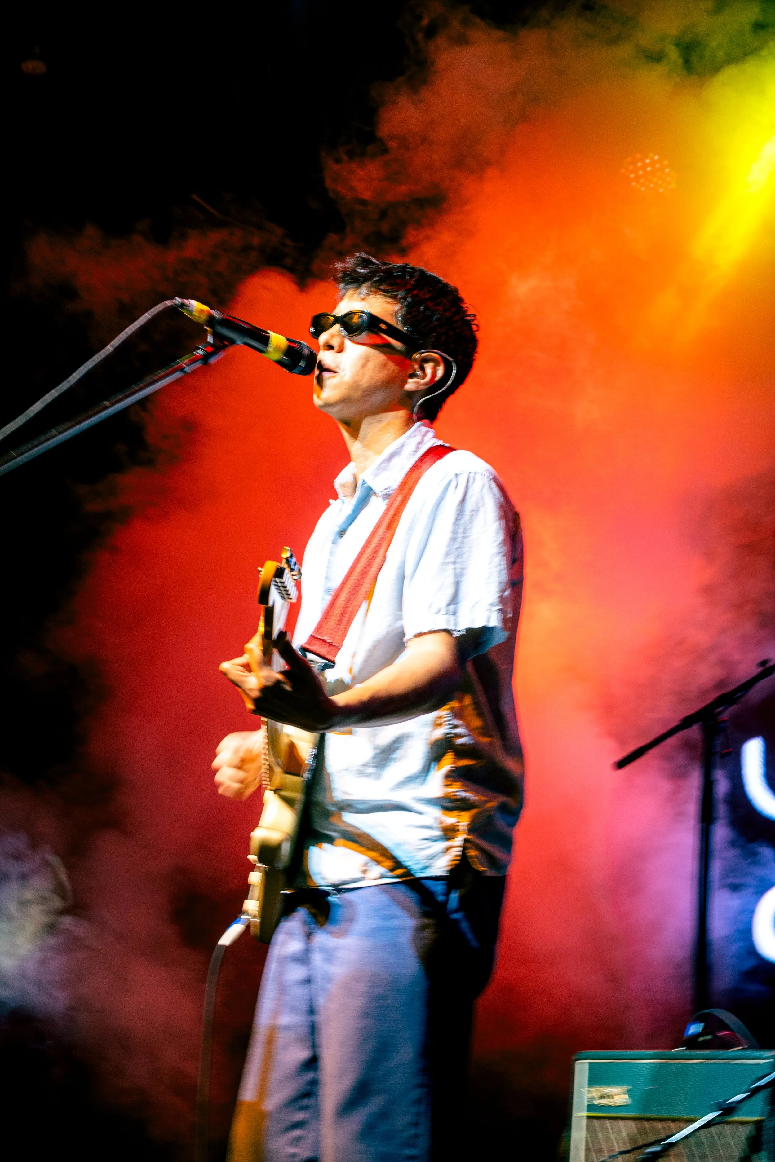 Joven músico tocando guitarra y cantando en un escenario con humo y luces de colores.