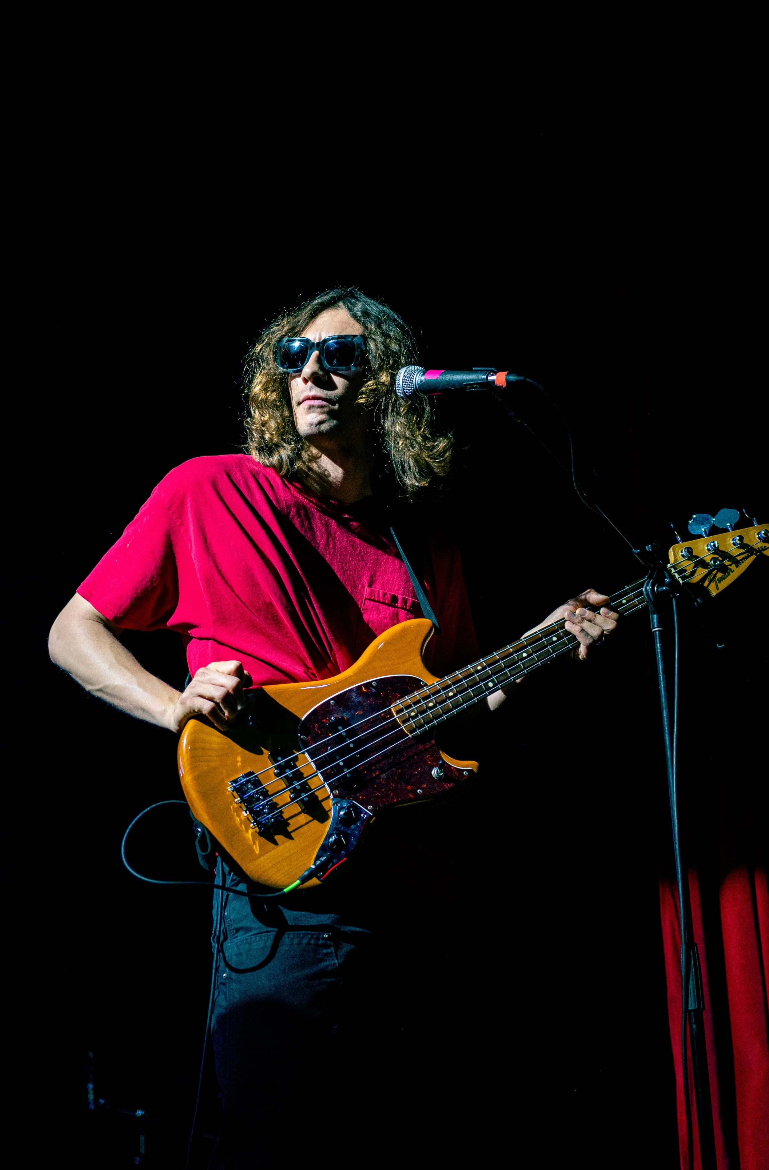 Un músico tocando la guitarra eléctrica en un escenario oscuro, usando gafas de sol y ropa de color rojo.