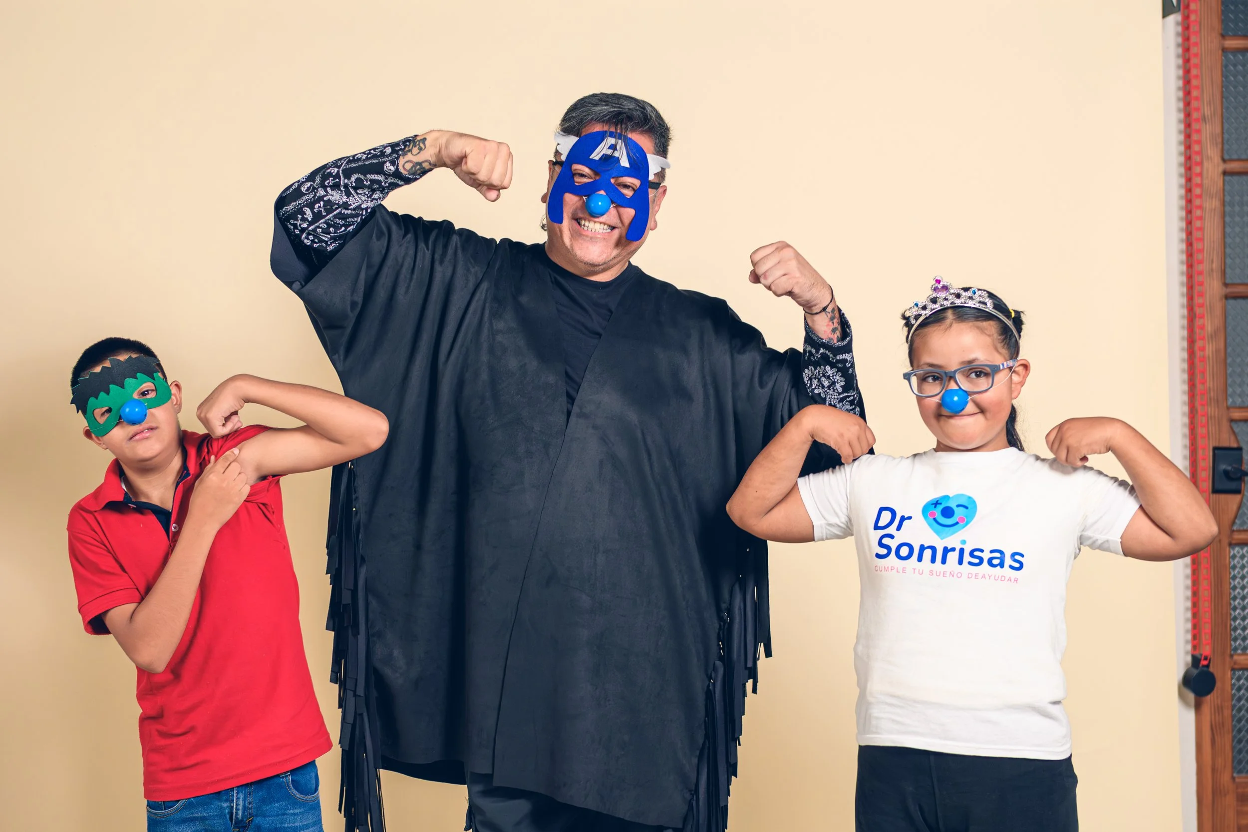Hombre y dos niños posando en un fondo claro, todos con narices y máscaras de superhéroes, haciendo músculos con los brazos. La mujer lleva una camiseta blanca con el logo de 'Dr Sonrisas' y los niños llevan narices y máscaras azules y verdes.