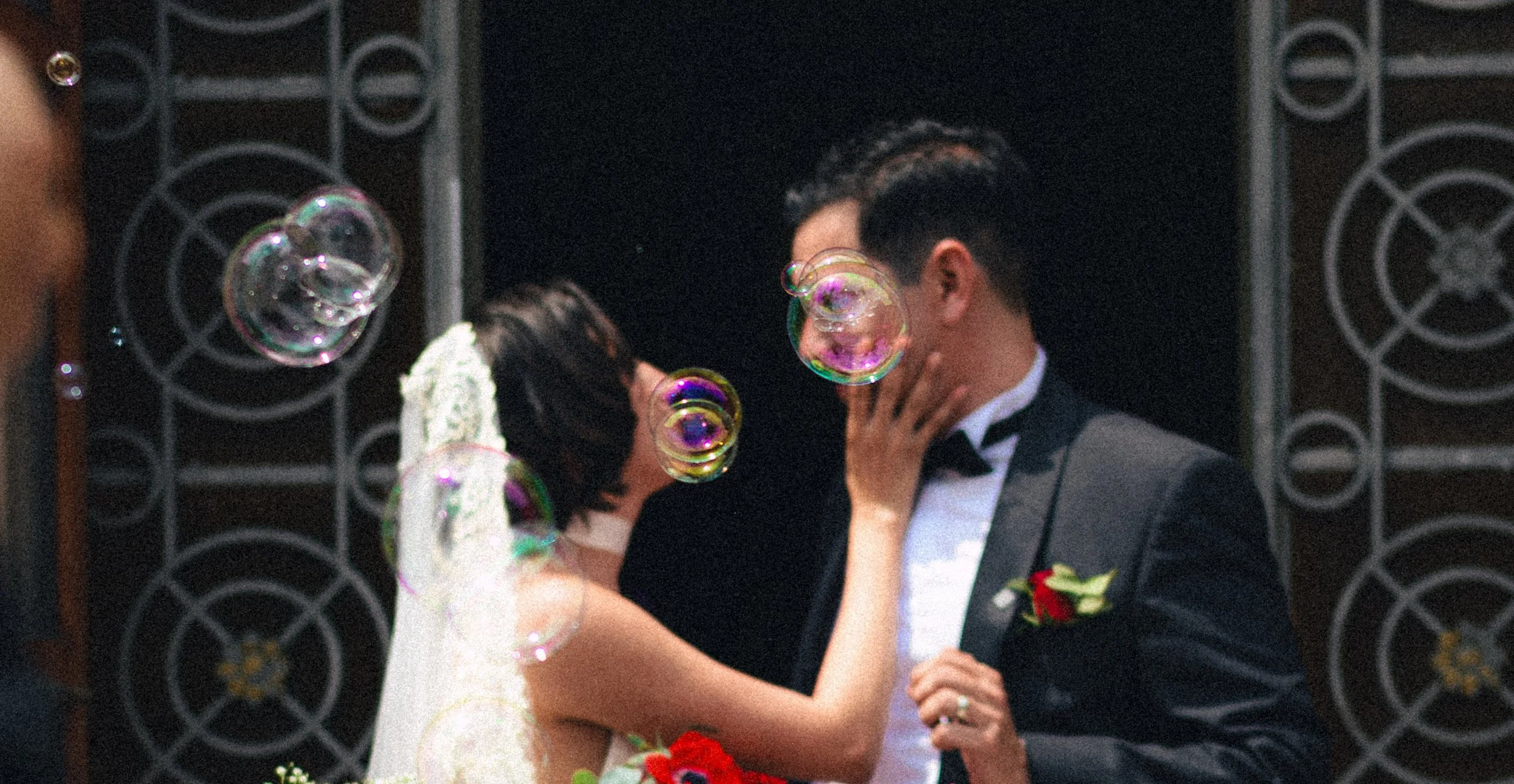 Una pareja en vestido de novia y traje de novio se besan en una boda, rodeados de burbujas de jabón y decoraciones negras en el fondo.