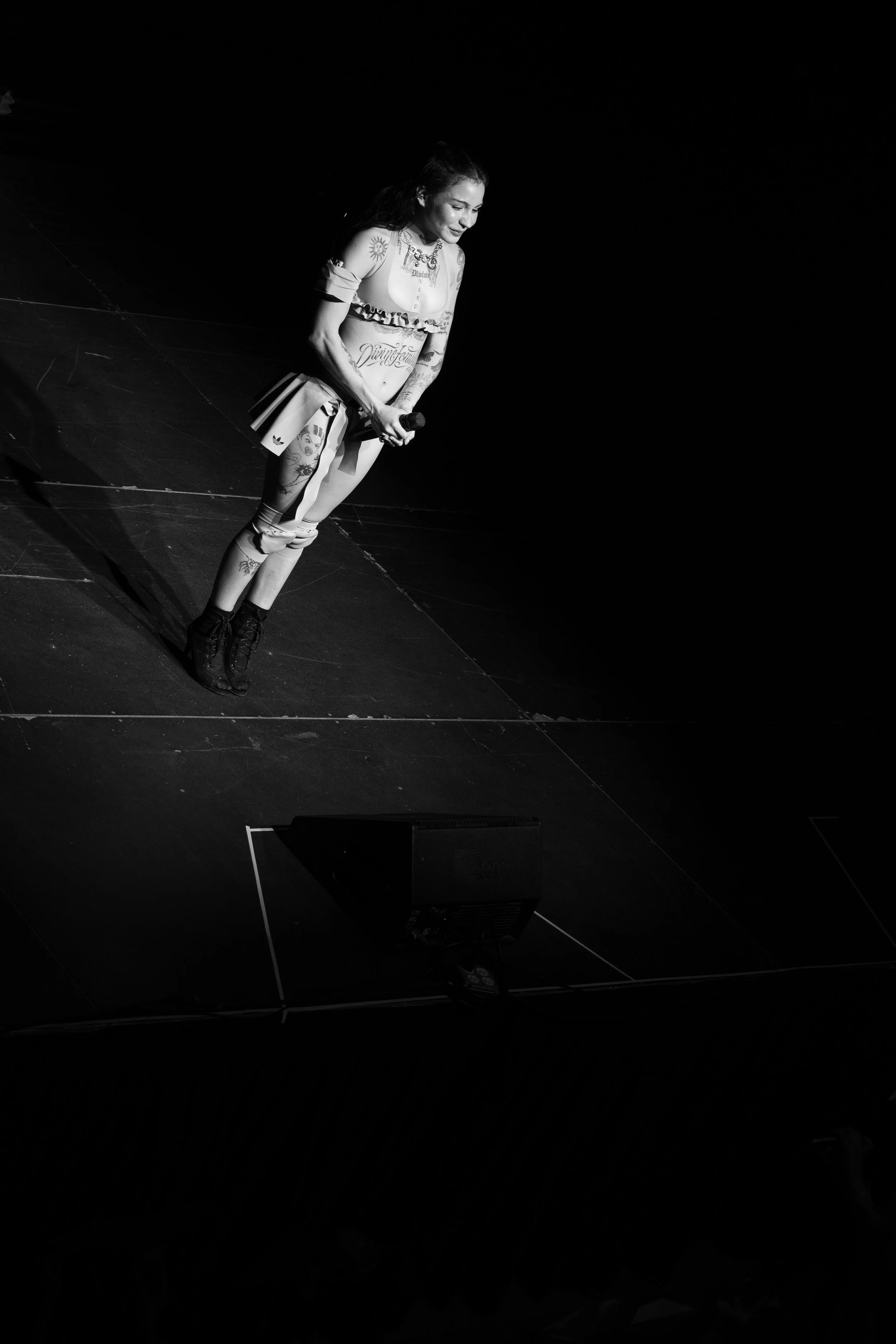 Una mujer con tatuajes en un escenario oscuro, sosteniendo un micrófono y sonriendo.
