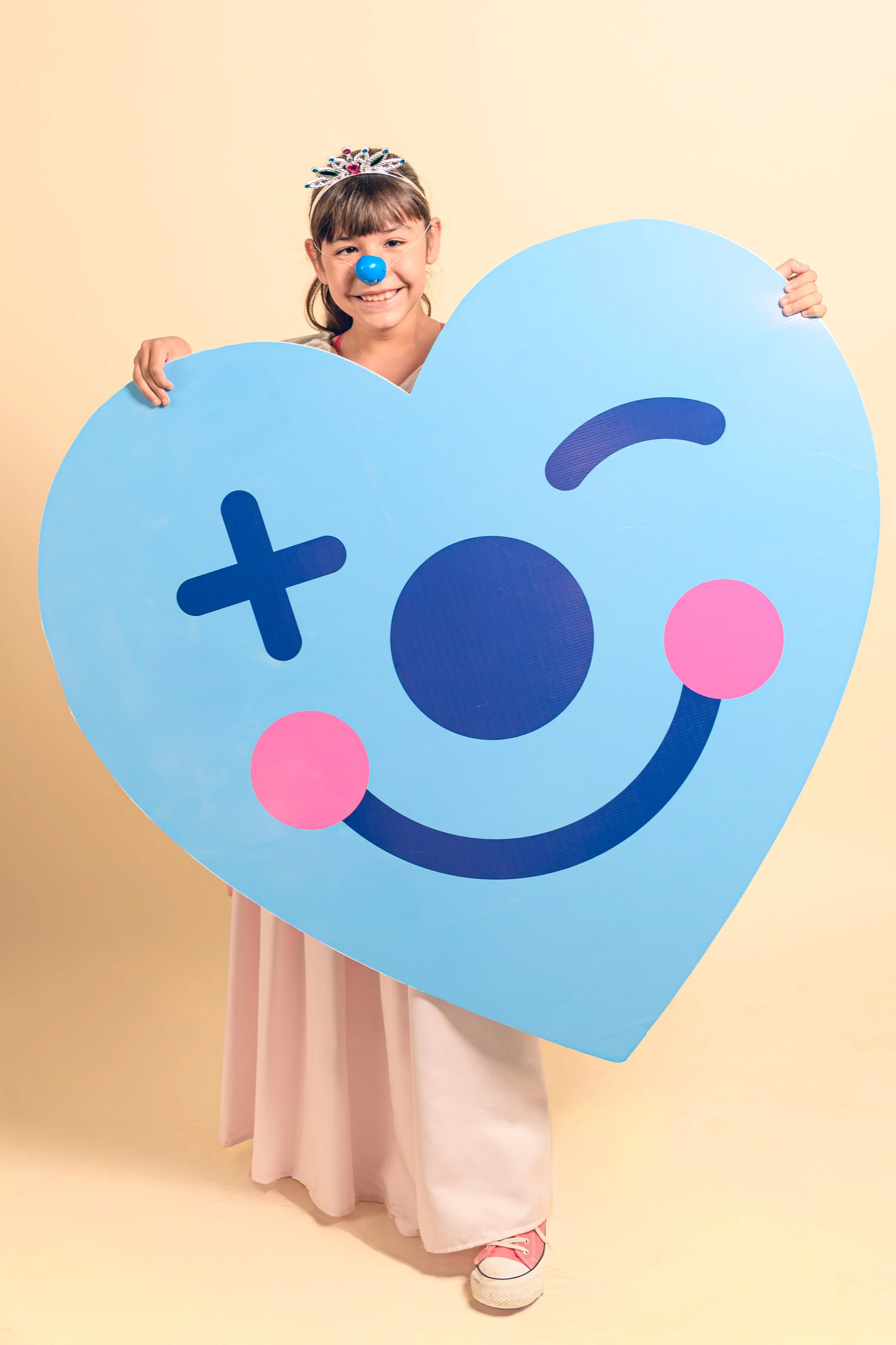 Niña sonriendo con una corona y nariz de payaso, sosteniendo un cartel grande con forma de corazón azul que tiene una carita sonriente y de un ojo cruzado.