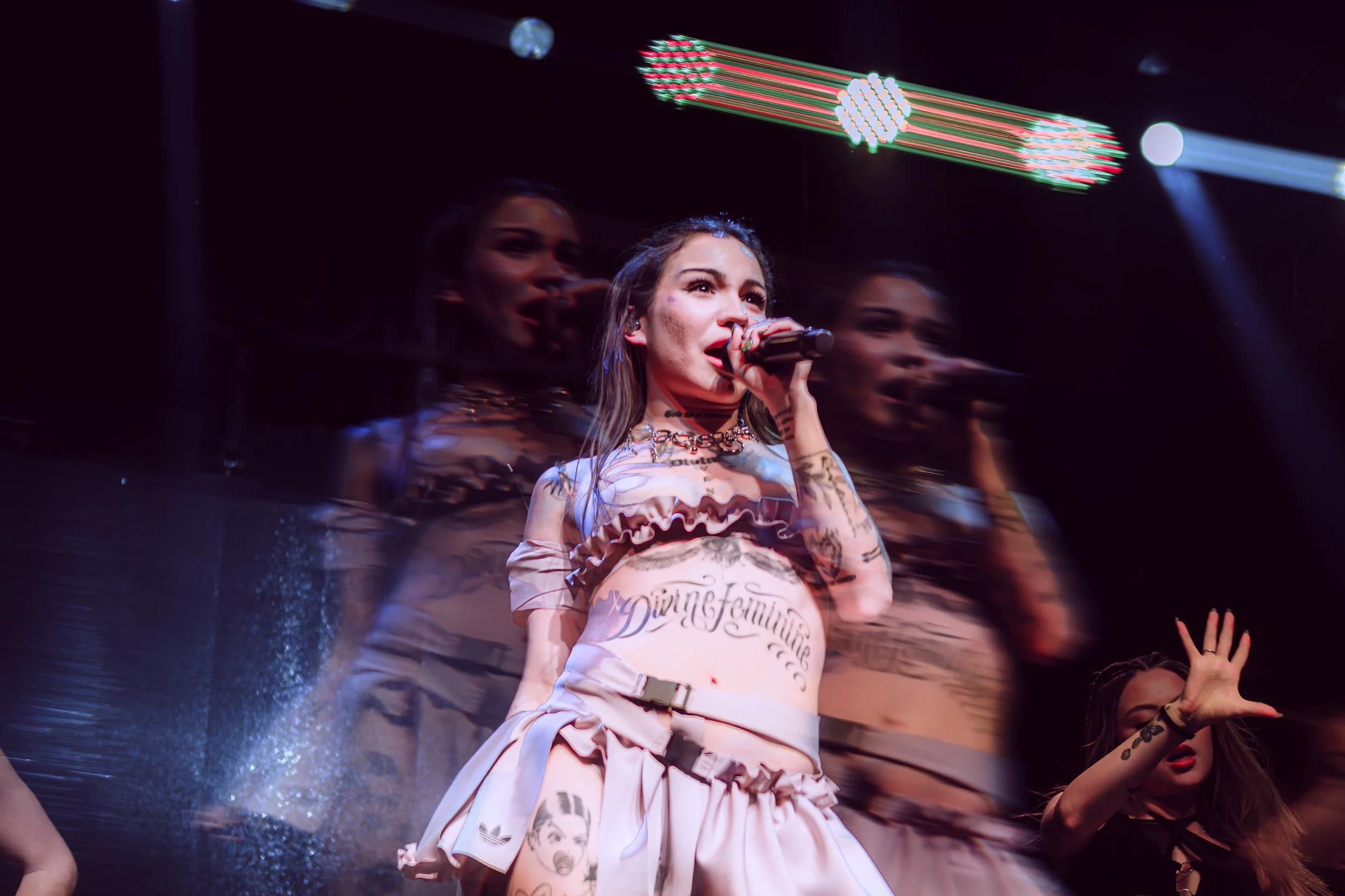 Una cantante en el escenario, rodeada de movimientos borrosos y luces de concierto, sosteniendo un micrófono y luciendo ropa con estampados y tatuajes visibles.