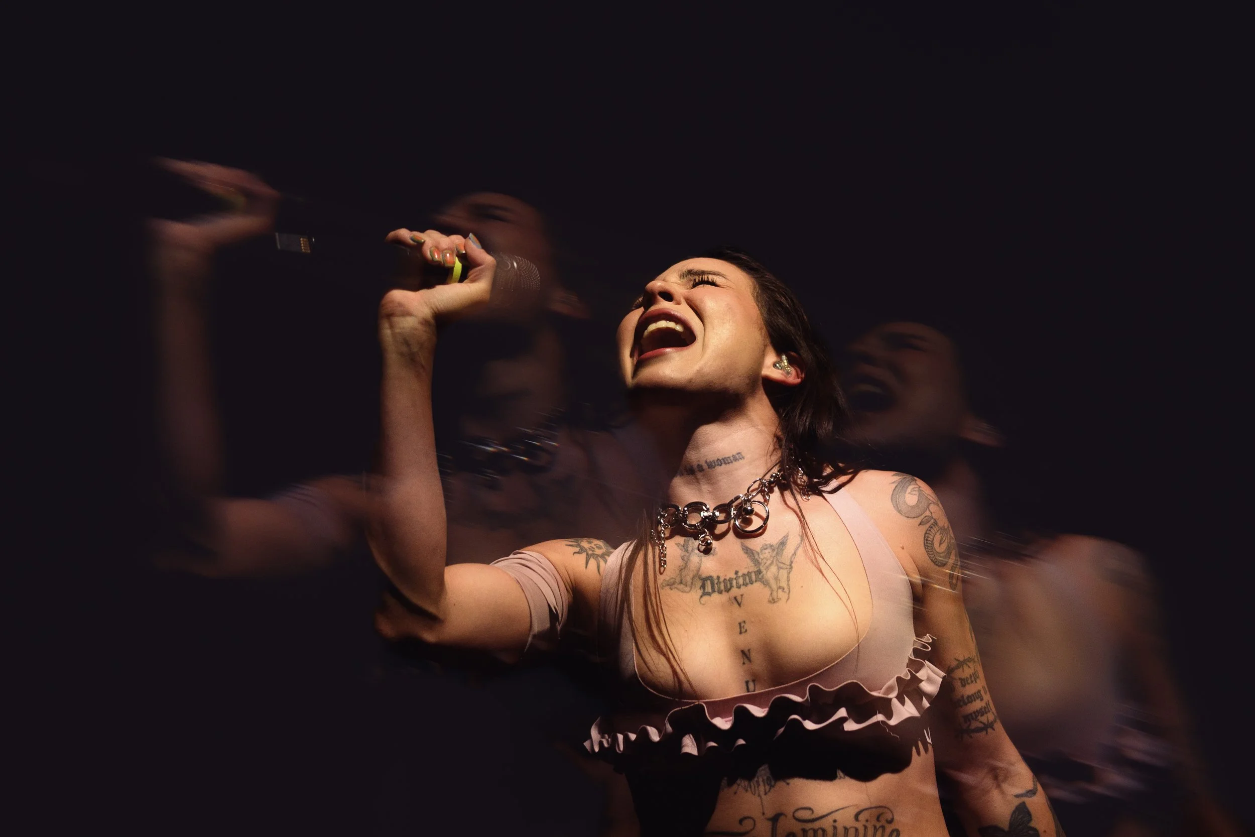 Una mujer con tatuajes, pelo largo y oscuro, canta con entusiasmo en un micrófono contra un fondo oscuro.
