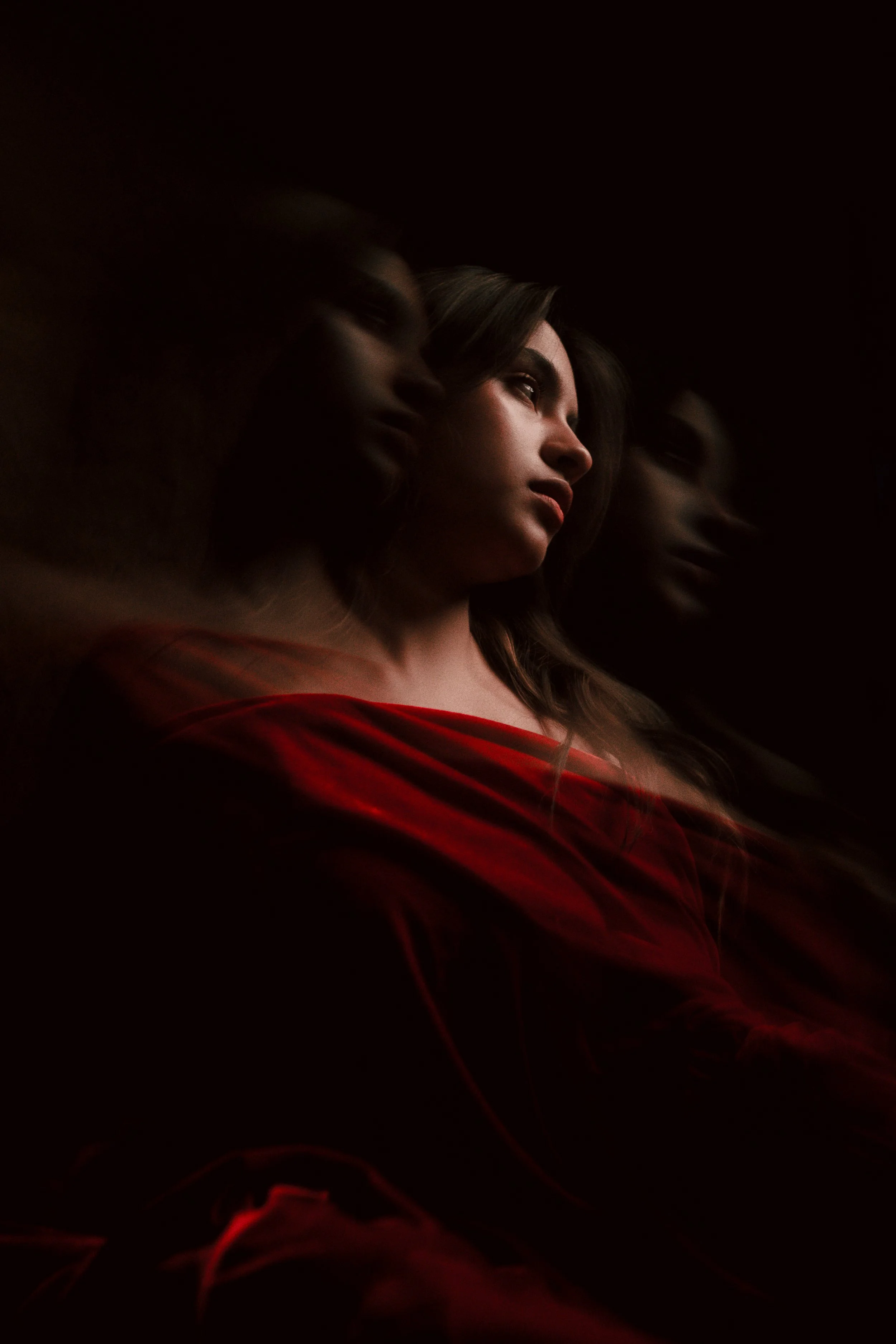 Una mujer con cabello largo y oscuro, vestida con ropa roja, mira pensativa hacia un lado en un fondo oscuro con reflejos de su rostro a la izquierda.