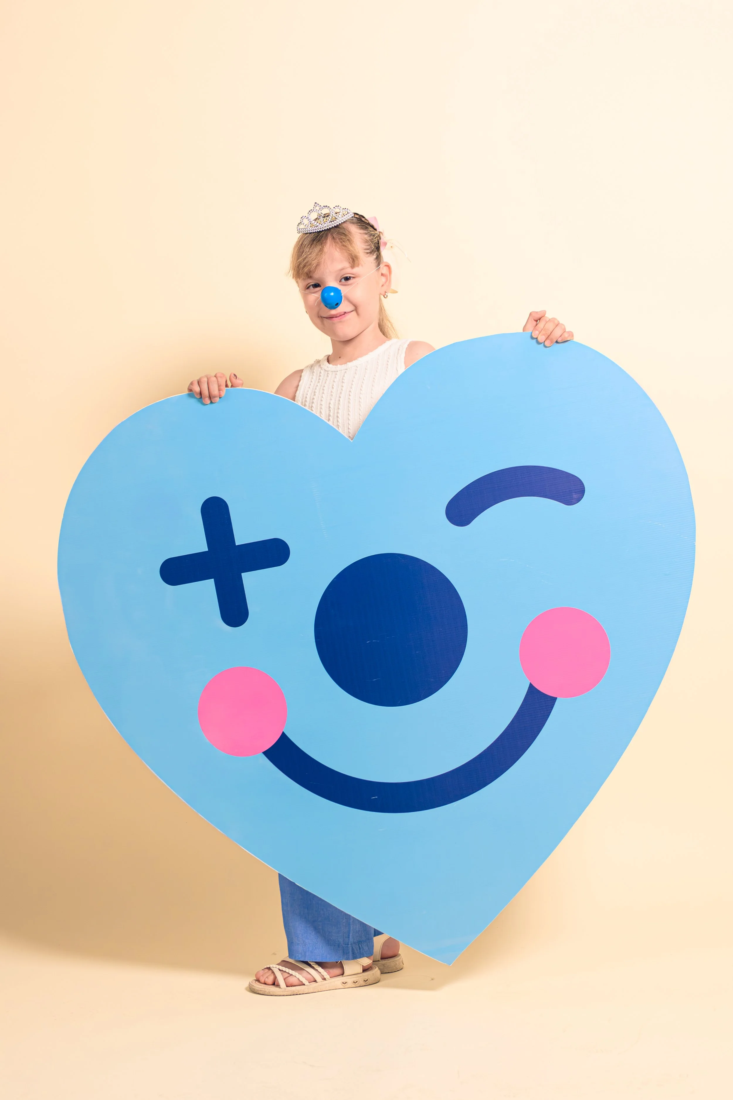 Niña con supuesta nariz de payaso, sosteniendo un cartel de corazón con cara sonriente, en fondo claro.