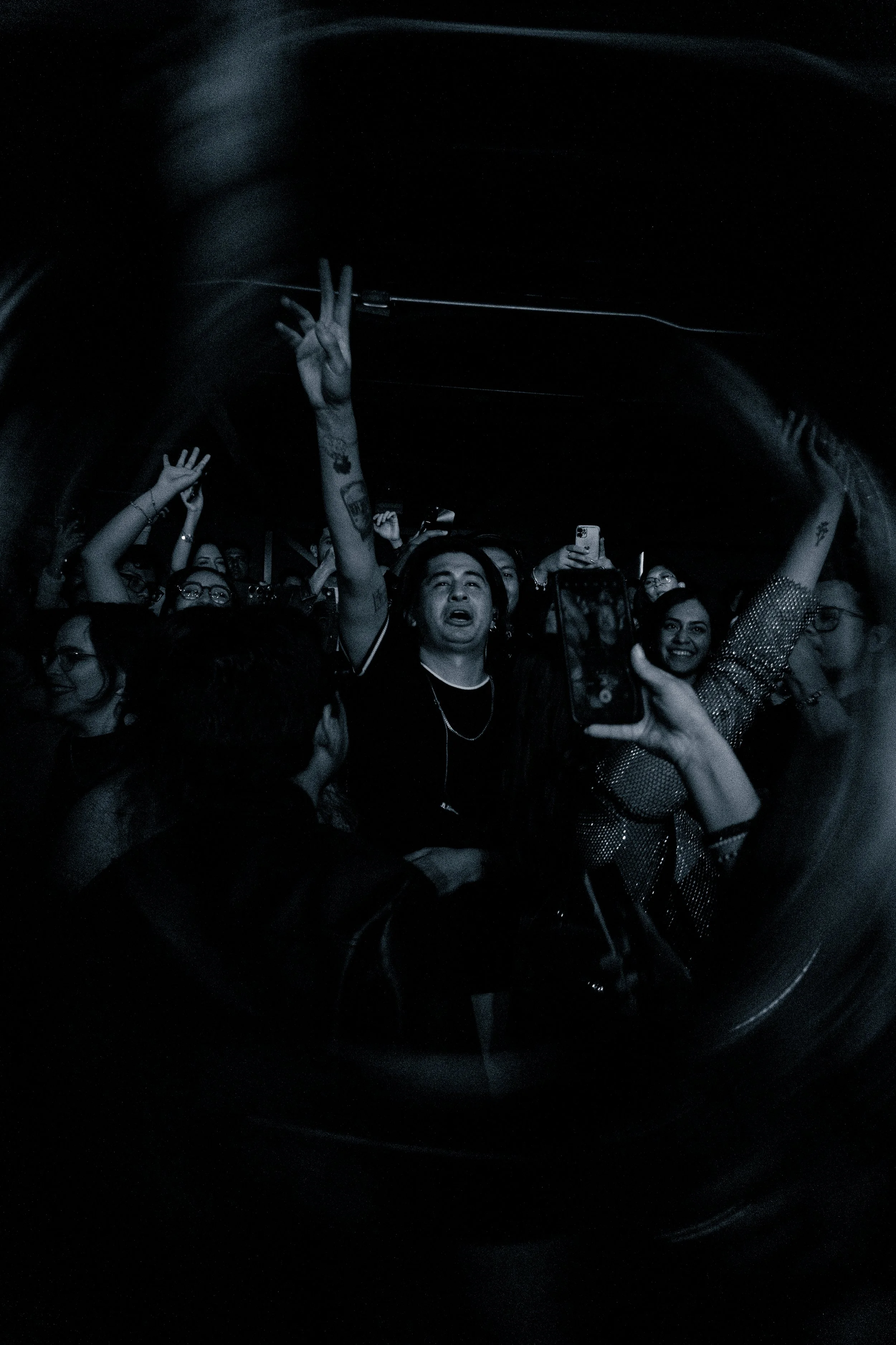 Grupo de personas en un concierto o evento, algunas con las manos en el aire y otras tomando fotos, en un ambiente oscuro.
