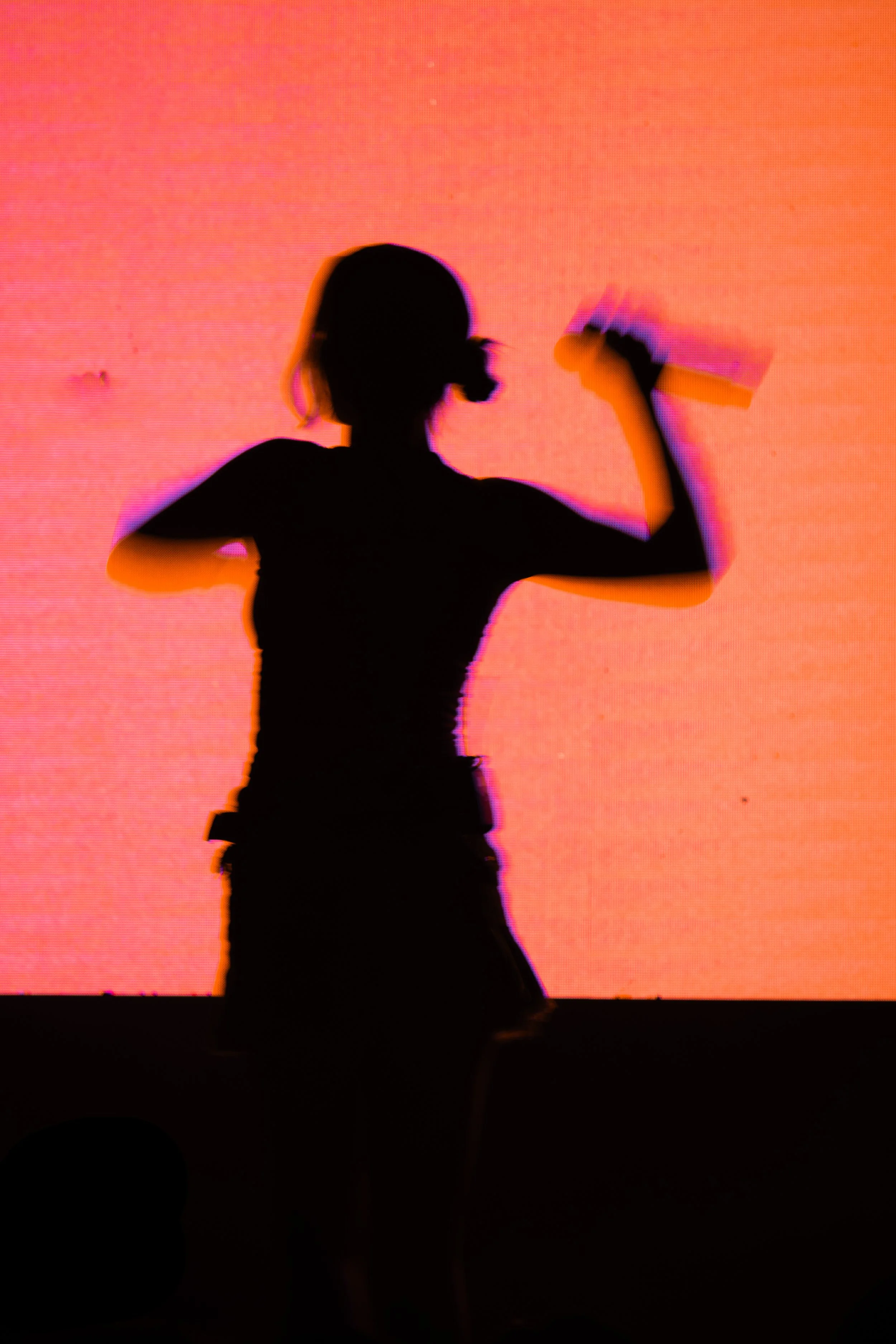 Silhouette de una niña con coletas, sosteniendo un libro o cuaderno en la mano derecha, mientras está en movimiento frente a una pantalla o fondo de color naranja.