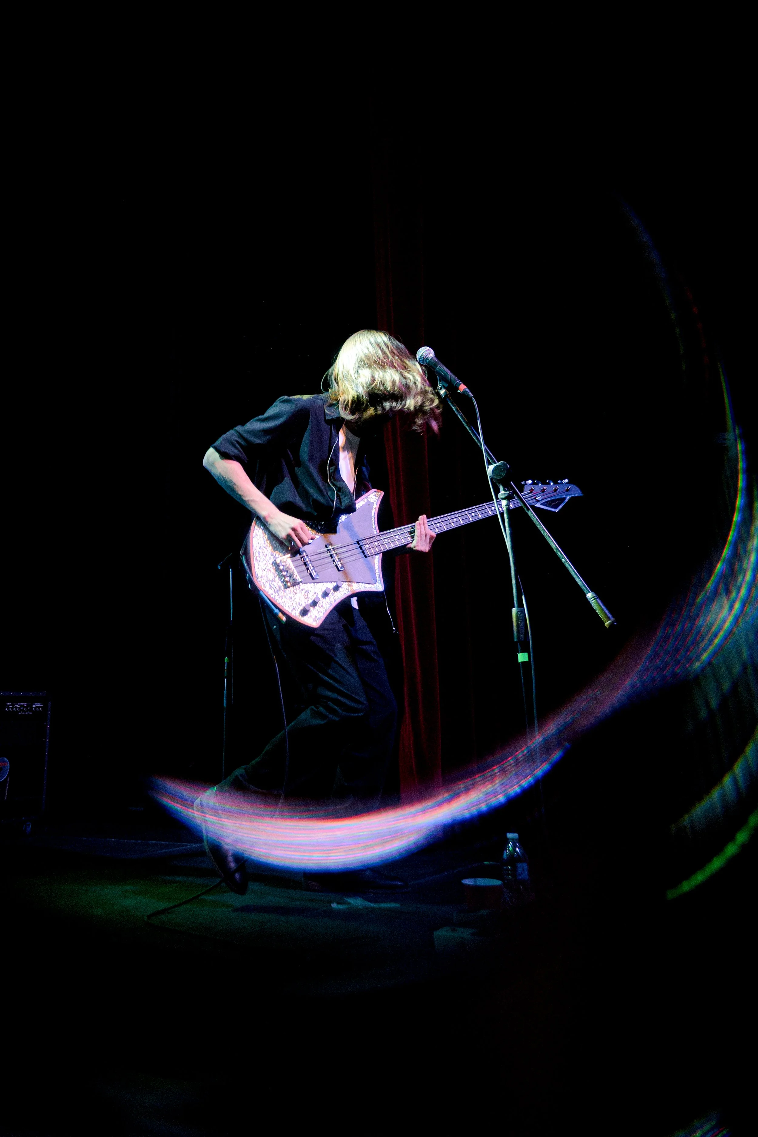 Músico tocando la guitarra en un escenario oscuro con un micrófono sobre un soporte y luces de escenario creando un efecto de movimiento alrededor.