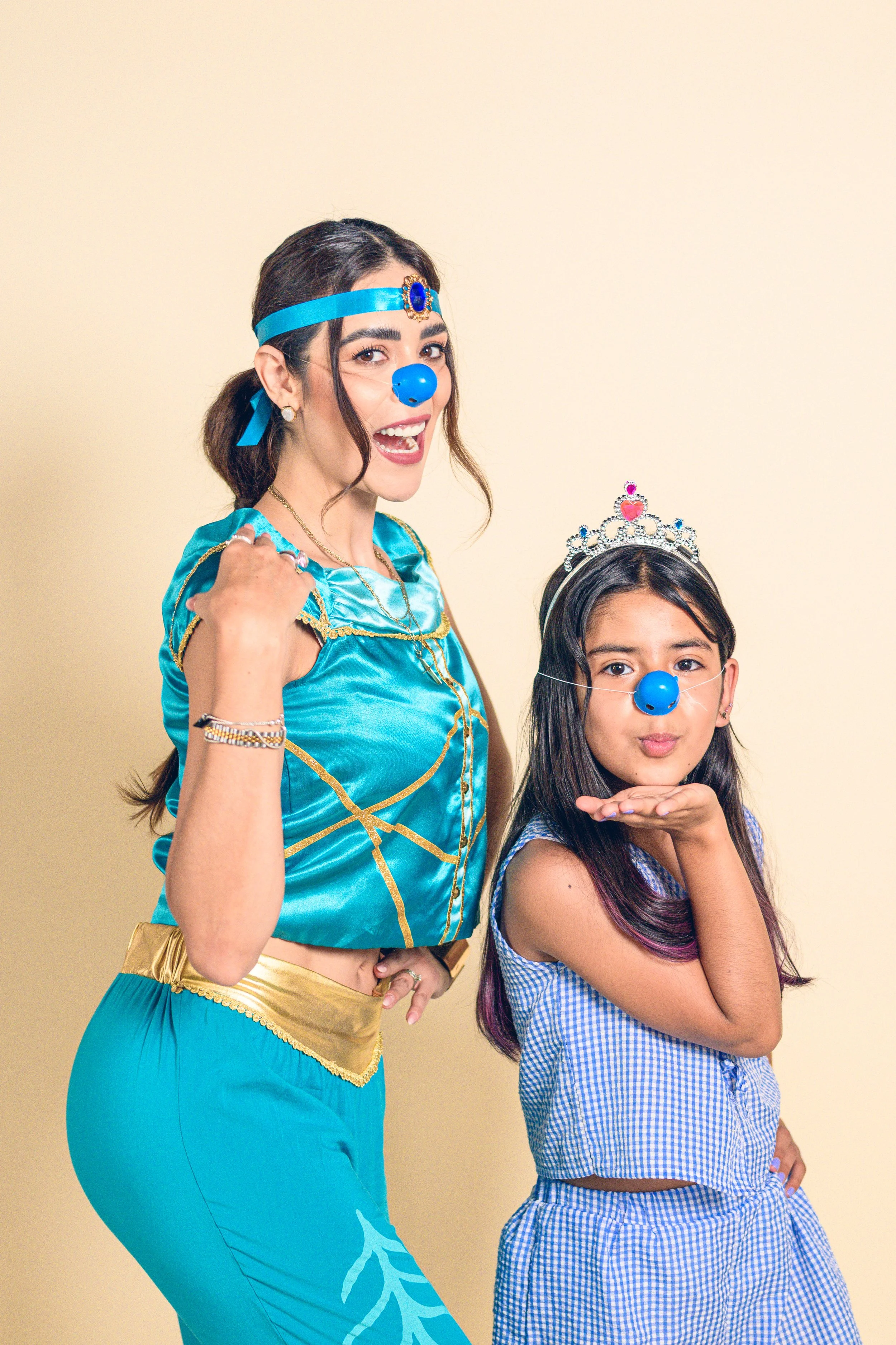 Dos mujeres con disfraces y narices azules, una vestida de princesa y otra con vestido a cuadros, posando juntas con fondo beige.