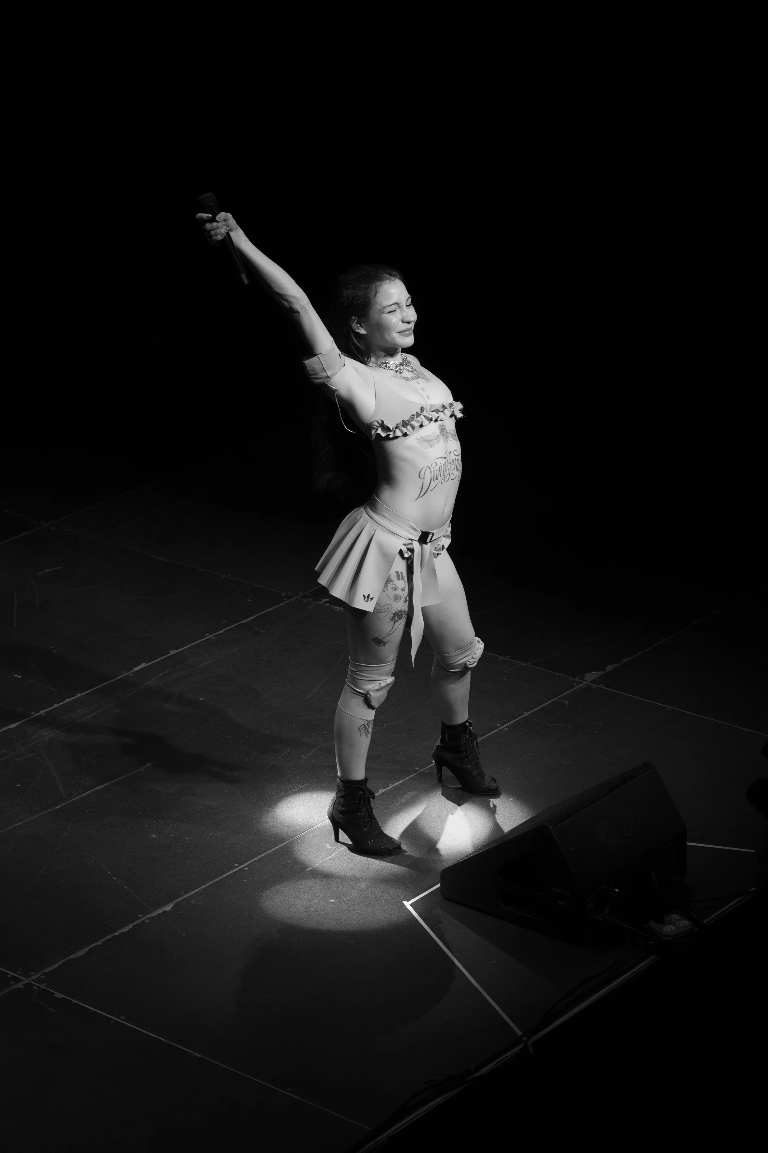 Una mujer en el escenario durante un espectáculo, sostiene un micrófono en su mano derecha, vestida con ropa llamativa y botas altas, en un fondo oscuro con un haz de luz sobre ella.