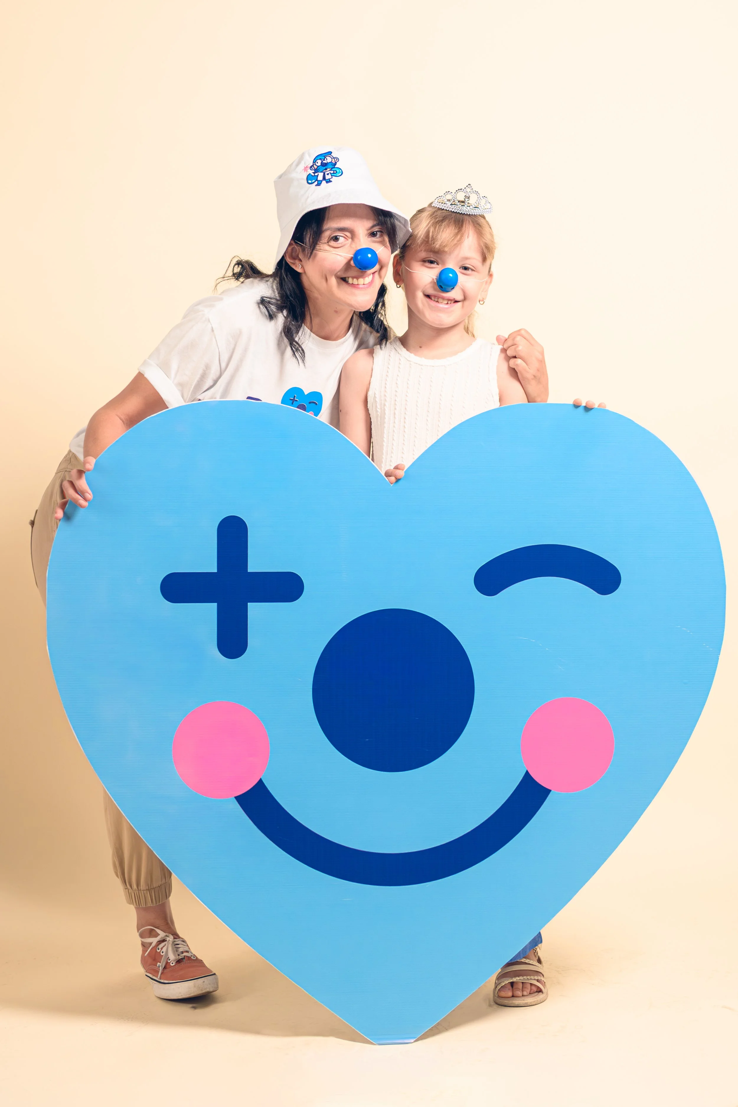 Dos personas, una adulta y una niña, posando con una gran figura de corazón azul con una cara sonriente y divertido, ambas con narices azules y accesorios blancos, en un fondo beige.