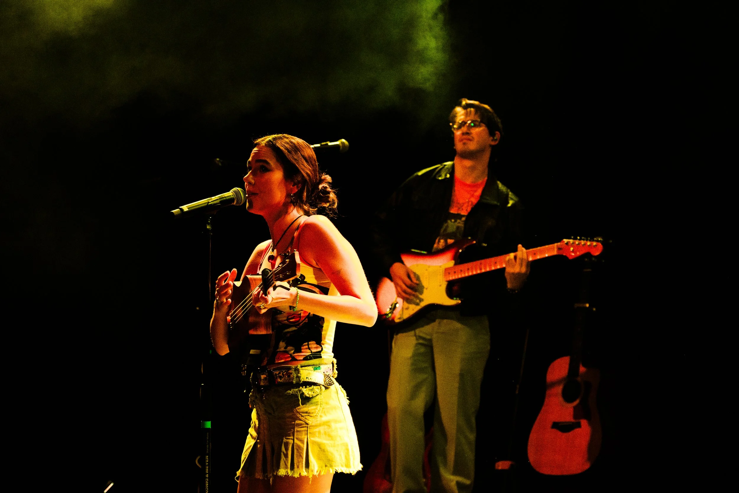 Dos músicos, una mujer tocando la guitarra y cantando frente a un micrófono, un hombre con gafas tocando la guitarra eléctrica en un escenario con poca iluminación