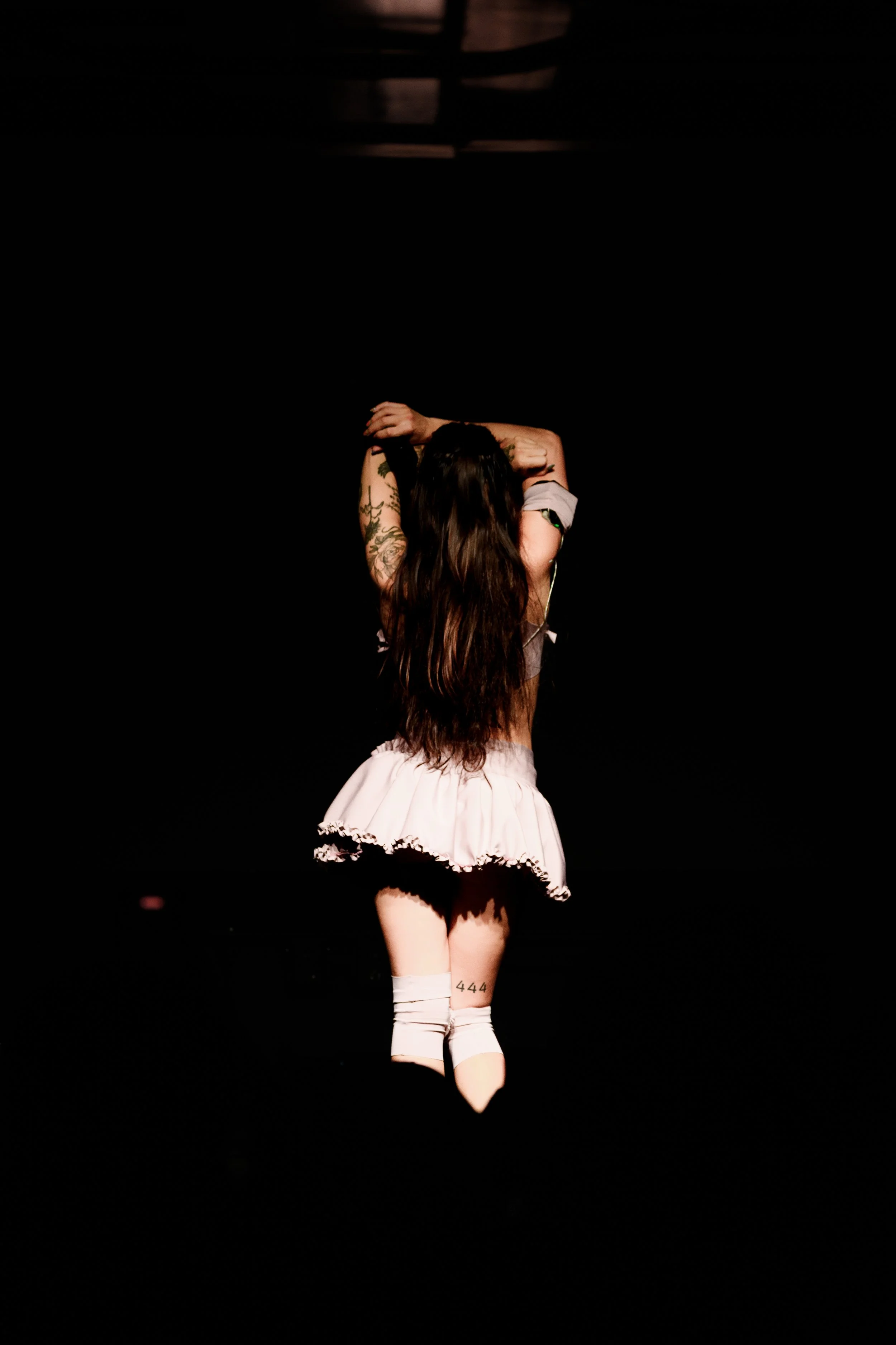 Una mujer con cabello largo y tatuajes, vestida con una falda rosa y calcetas blancas, posa en una escena con fondo negro, con las manos en la cabeza.