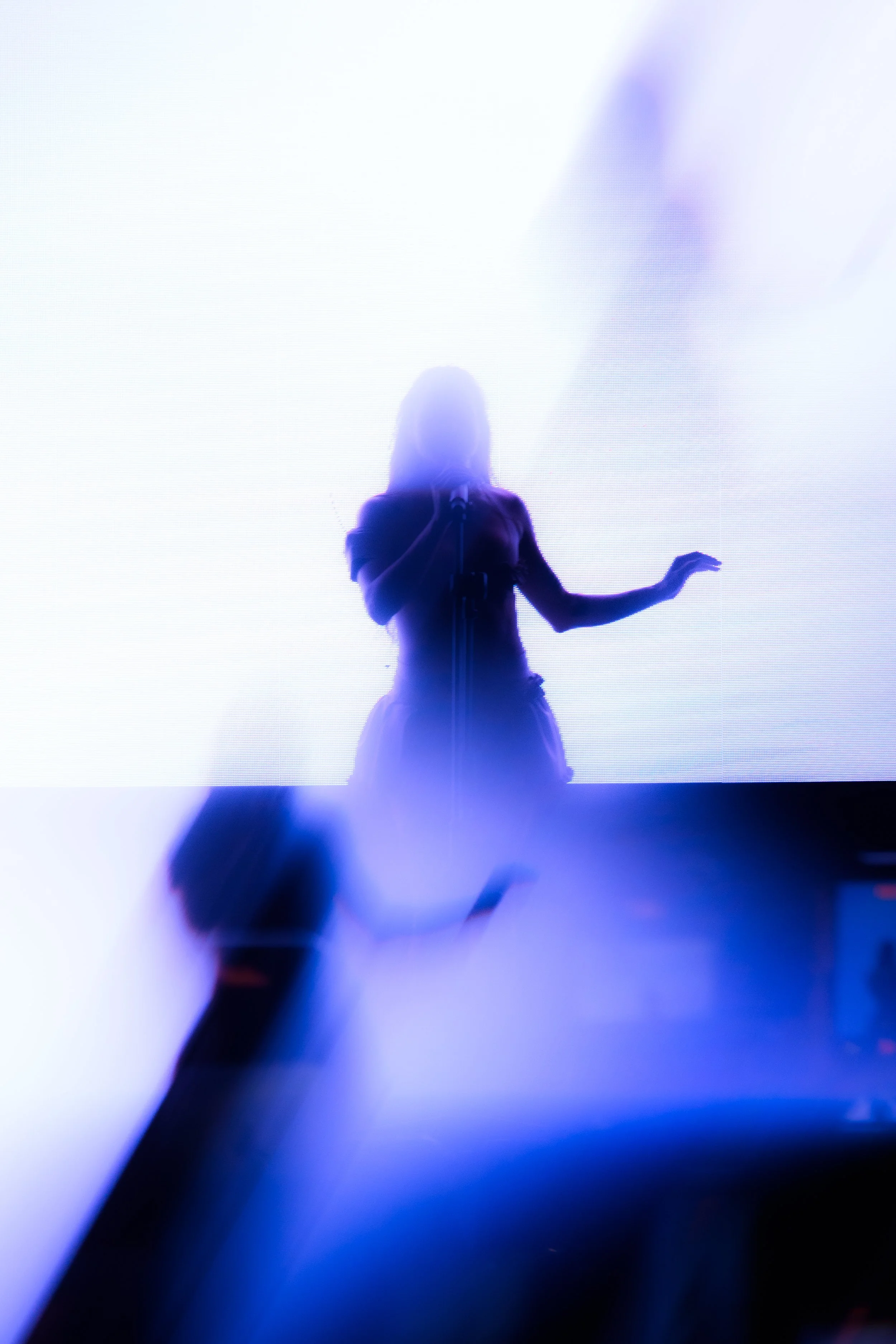 Silhouette de una mujer cantando o hablando en un escenario con fondo brillante. Ciudadano en actitud expresiva, con luz de fondo azul y blanco.