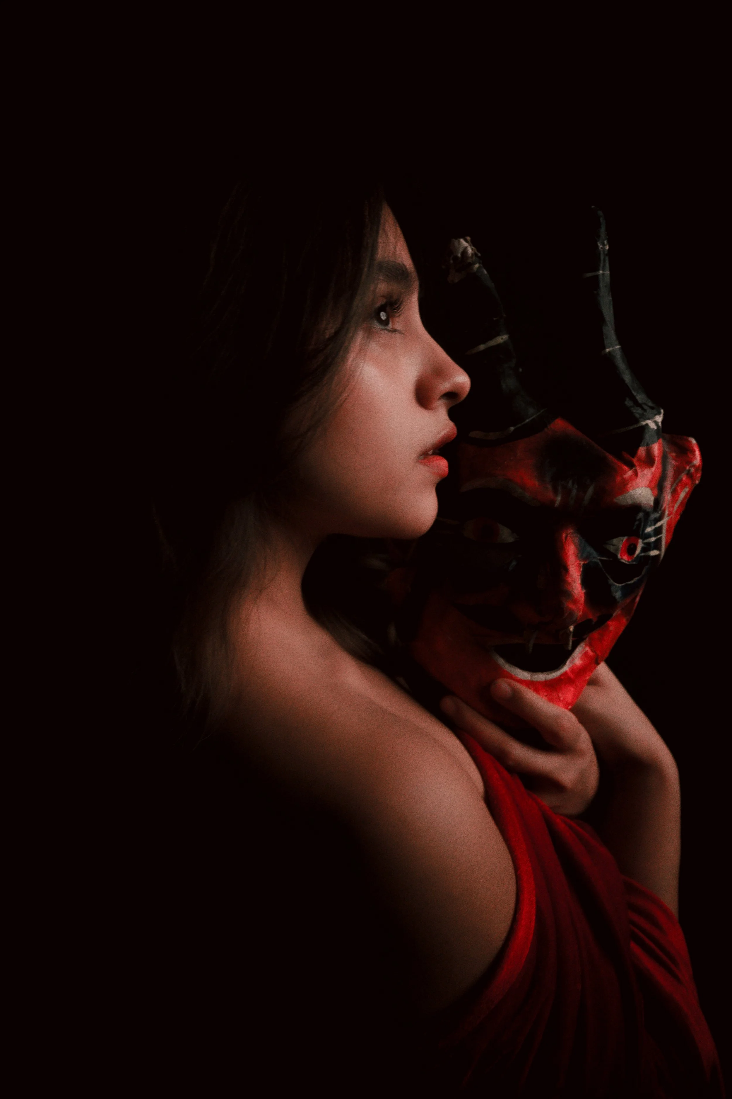 Mujer con piel clara sosteniendo una máscara de madera color rojo y negro, con características enojadas y ojos rojos, fondo oscuro.
