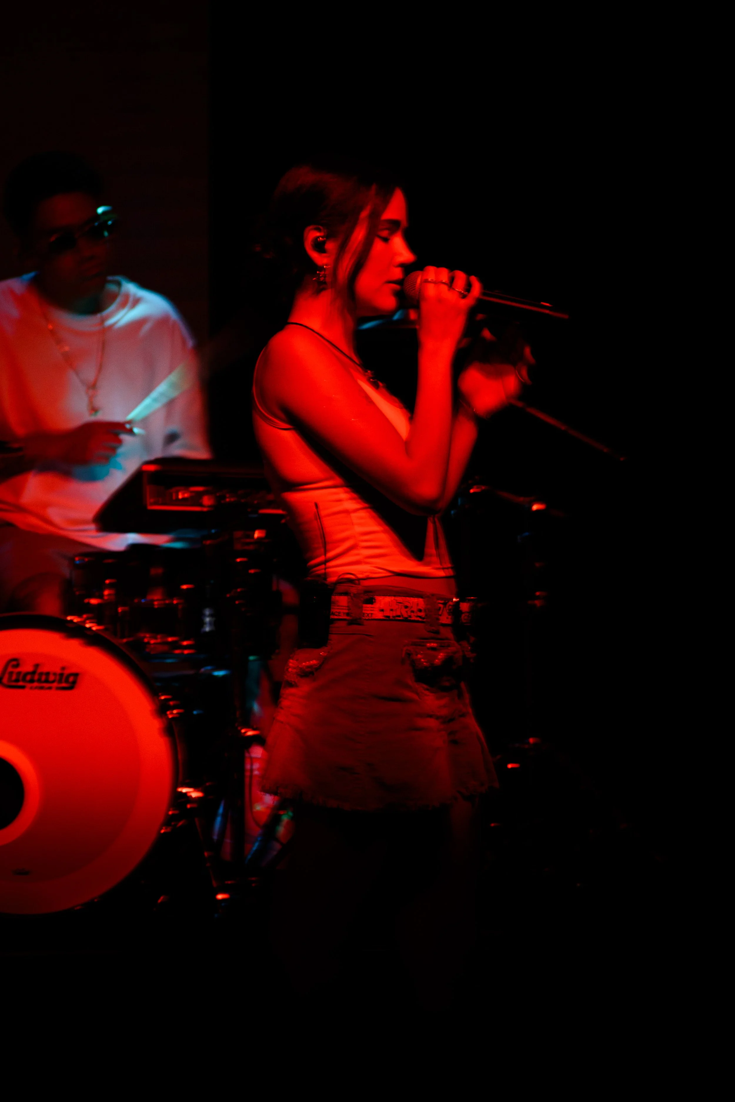 Una mujer cantando con micrófono en un escenario con iluminación roja, acompañada por un músico con instrumentos en un fondo oscuro.