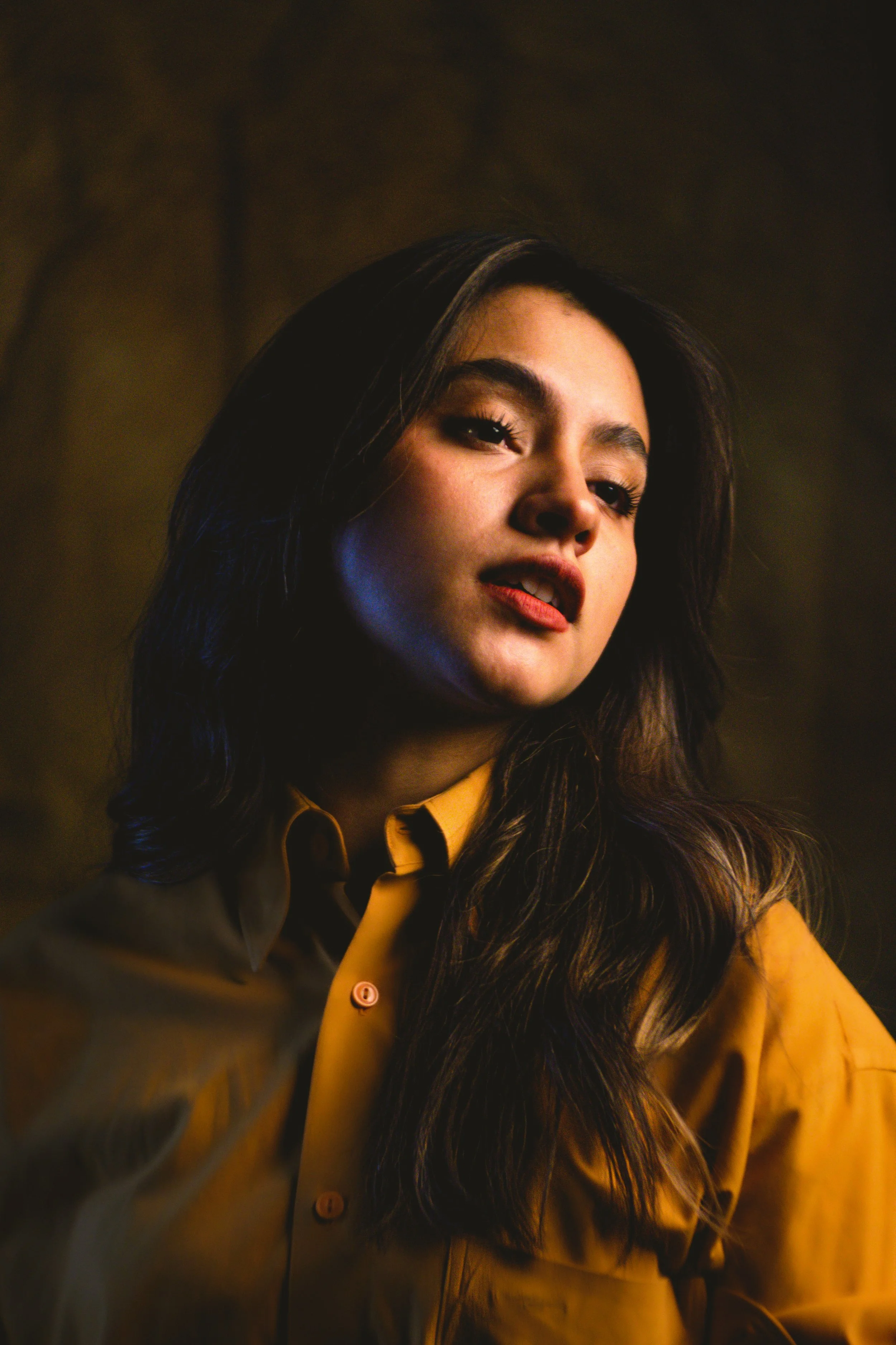 Retrato de una mujer joven con cabello oscuro y largo, vestida con camisa amarilla, en un fondo oscuro.