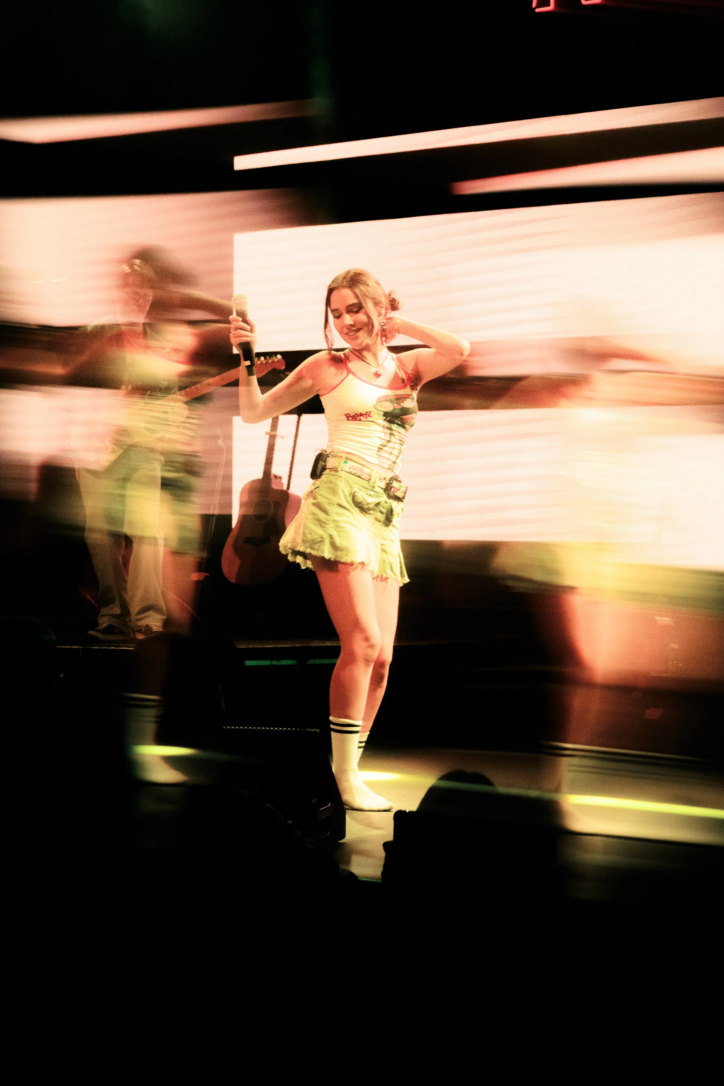 Una joven mujer bailando en un escenario con un micrófono, con música y luces de fondo, rodeada de otras personas que también parecen ser músicos o bailarines.