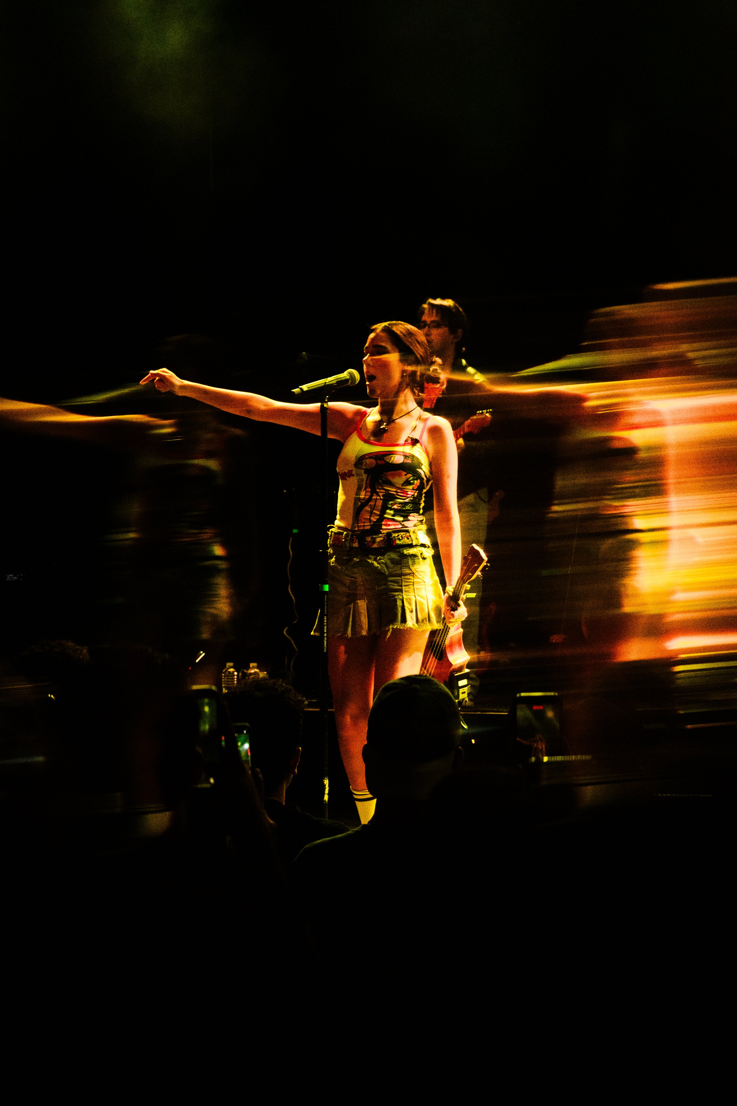 Una mujer cantando en un concierto con un micrófono, con una guitarra en la espalda, acompañada por una banda en el escenario con luces y efectos de movimiento.