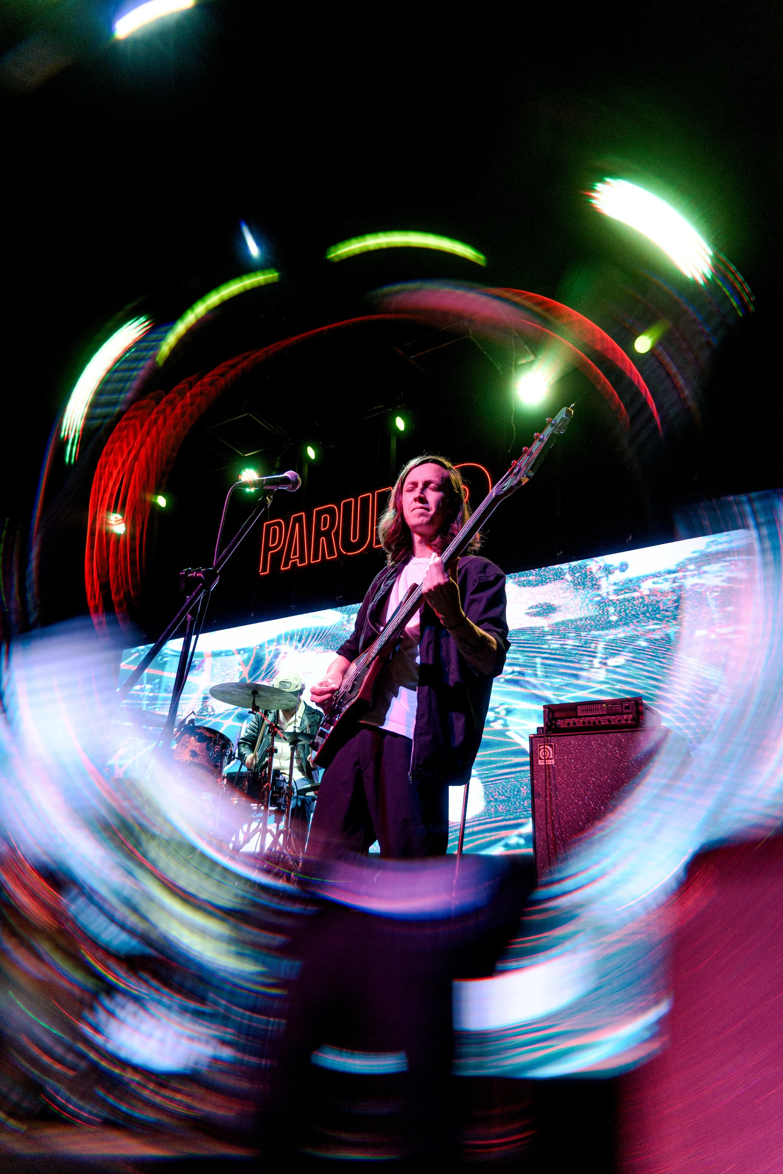 Un músico tocando la guitarra eléctrica en un escenario con luces de colores y una pantalla detrás que dice 'PARU'.