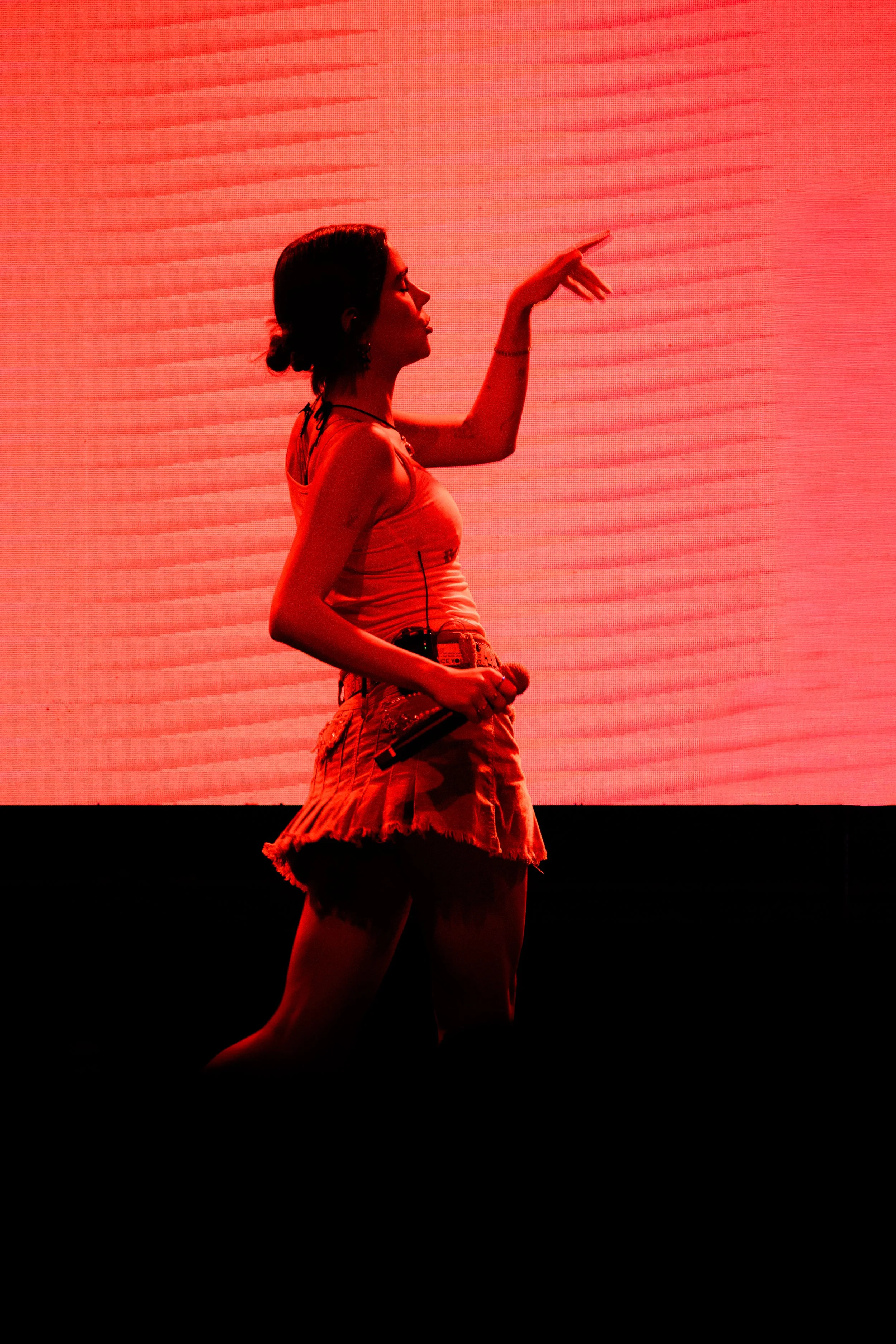 Mujer en escenario con fondo rojo, sosteniendo micrófono en la mano, haciendo un gesto con la otra mano.