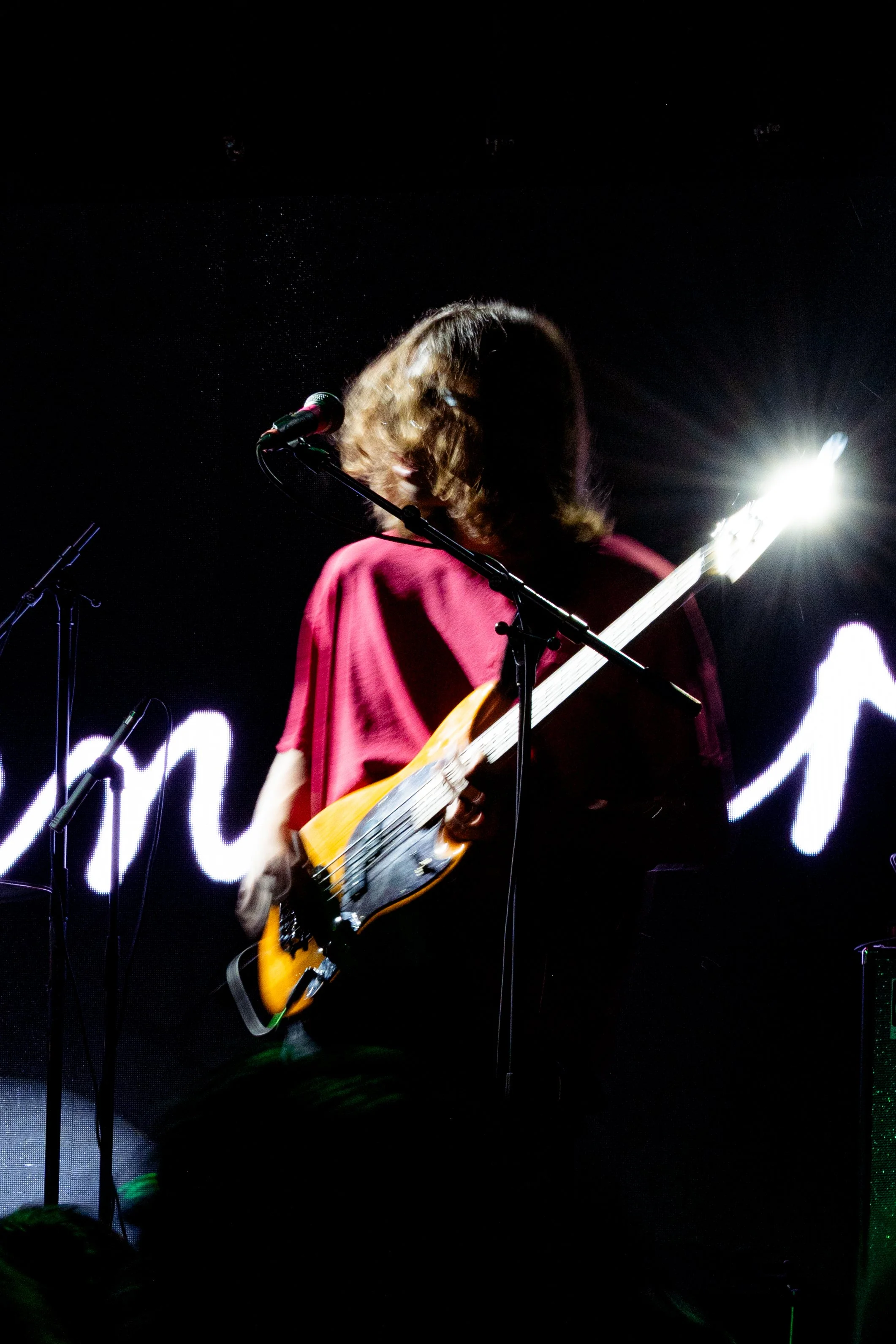 Persona tocando guitarra eléctrica en un escenario con fondo iluminado y luz brillante.