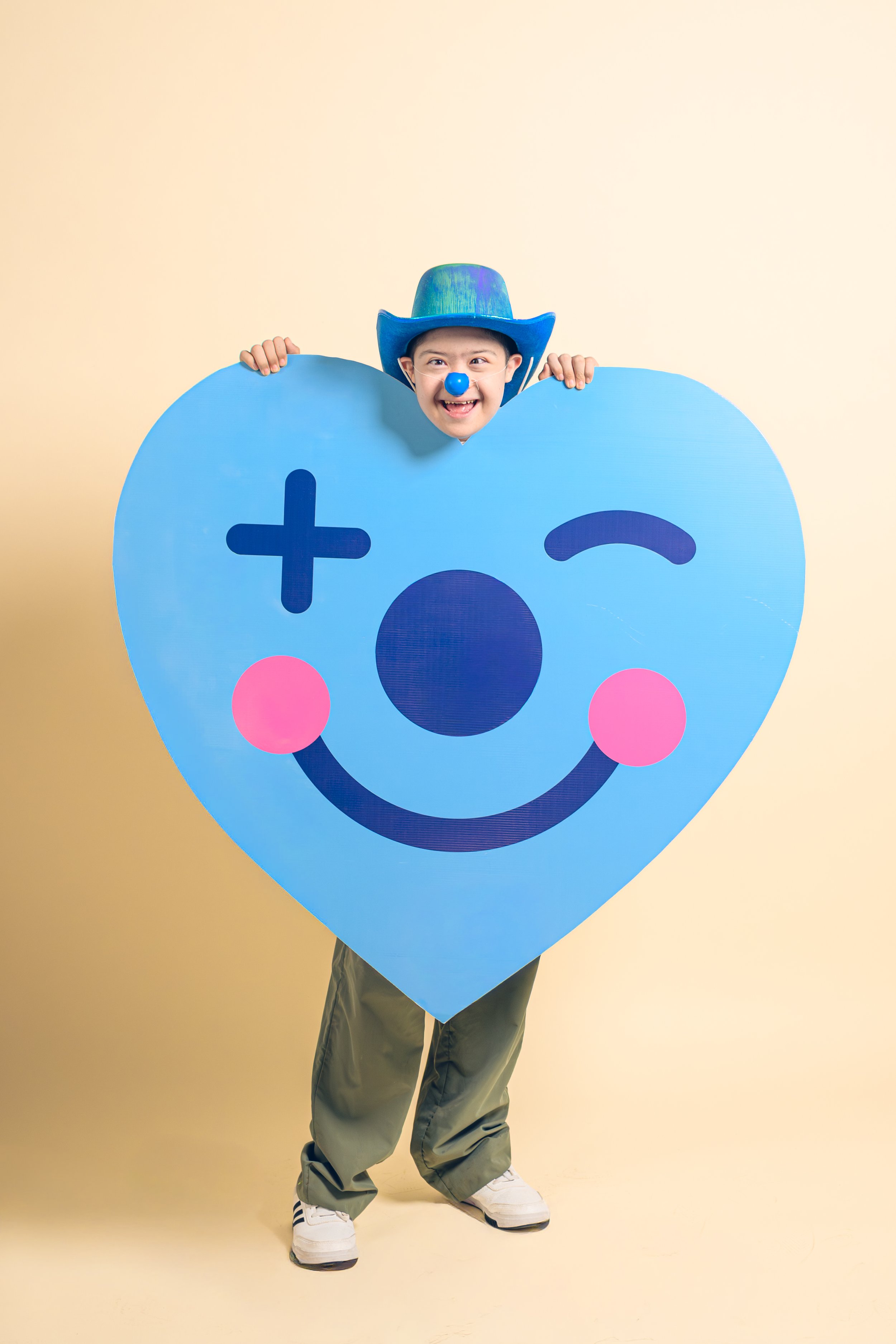 Niño disfrazado con un sombrero azul, nariz azul, y maquillaje, sosteniendo un corazón grande con una cara sonriente y un ojo cerrado, en un fondo beige.