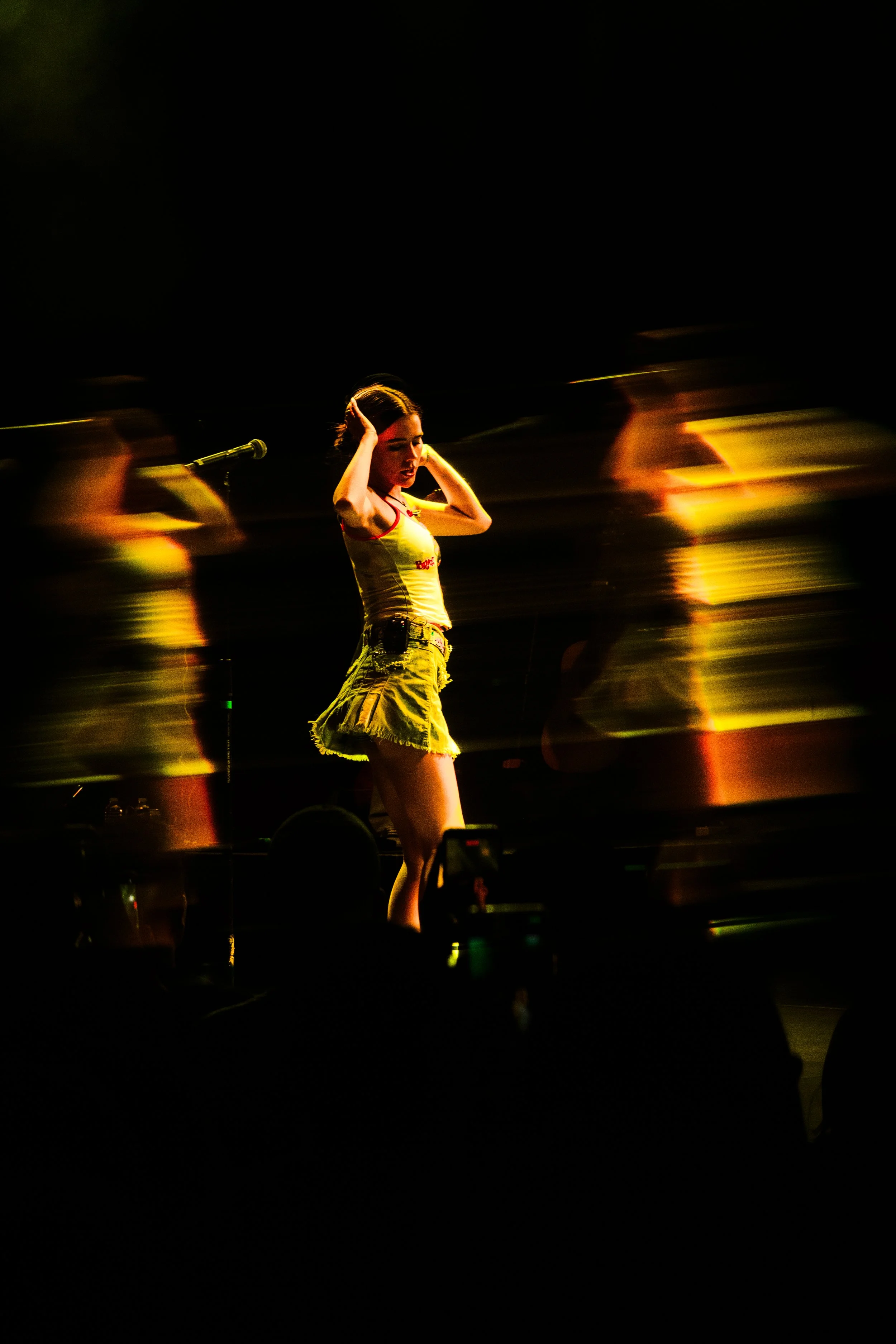 Una mujer en un escenario de concierto ajustándose el cabello mientras canta, acompañada por un grupo de personas que aparecen borrosas y en movimiento, con un fondo oscuro.