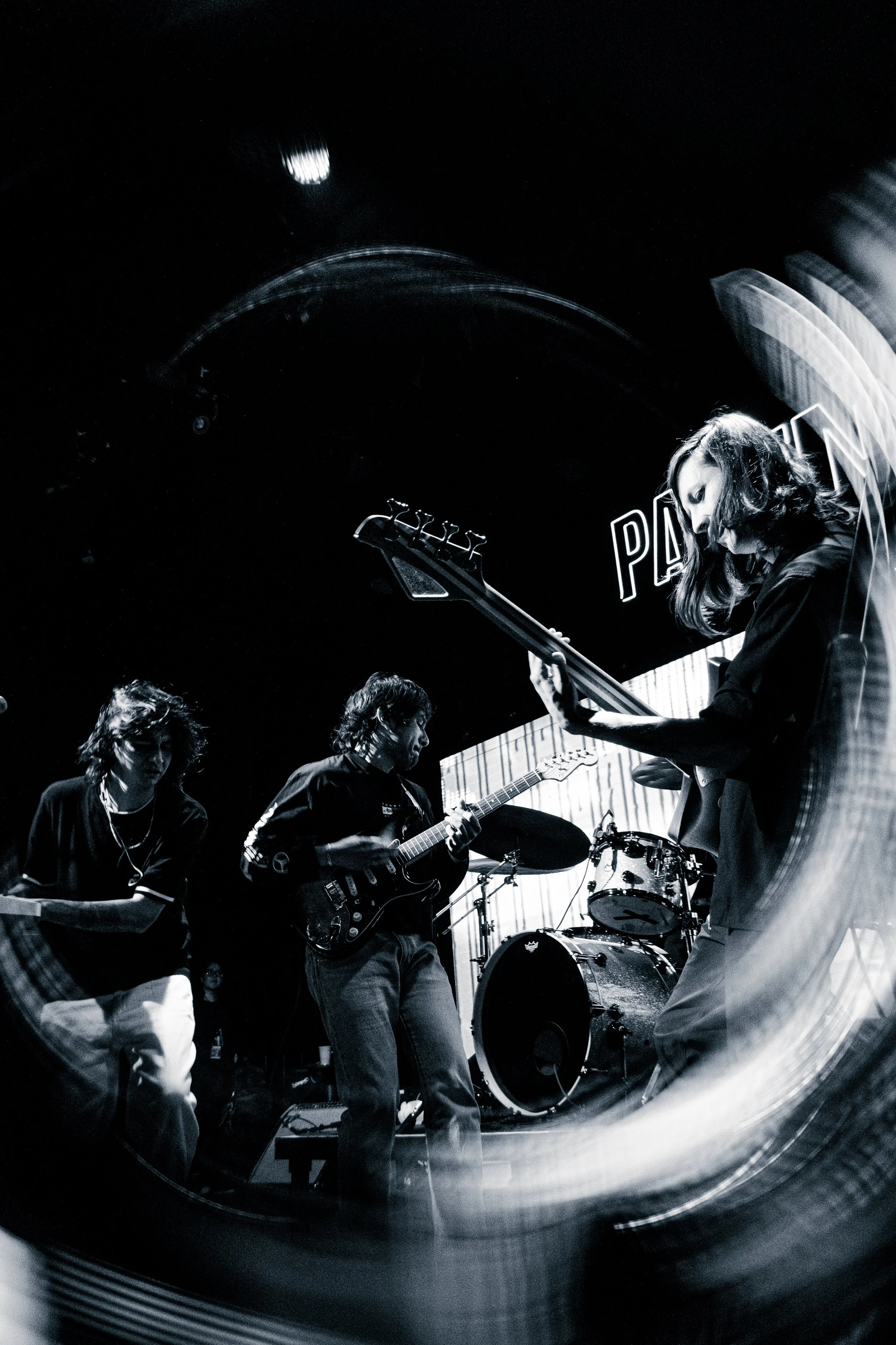 Miembros de una banda de música tocando en vivo en un escenario, con instrumentos de cuerda y batería, en un ambiente oscuro y dinámico.