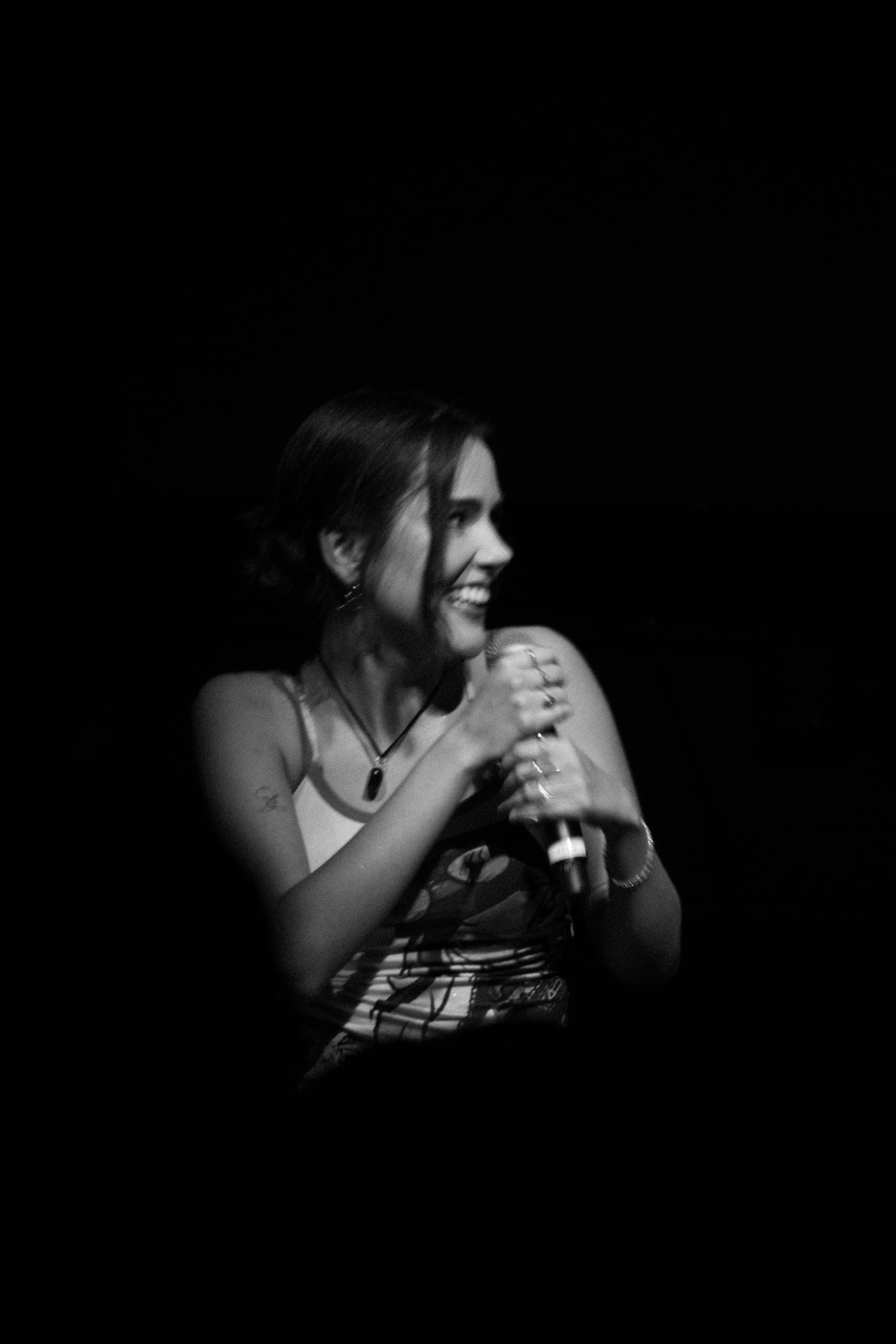 Mujer cantante con micrófono, sonriendo, en un escenario oscuro.