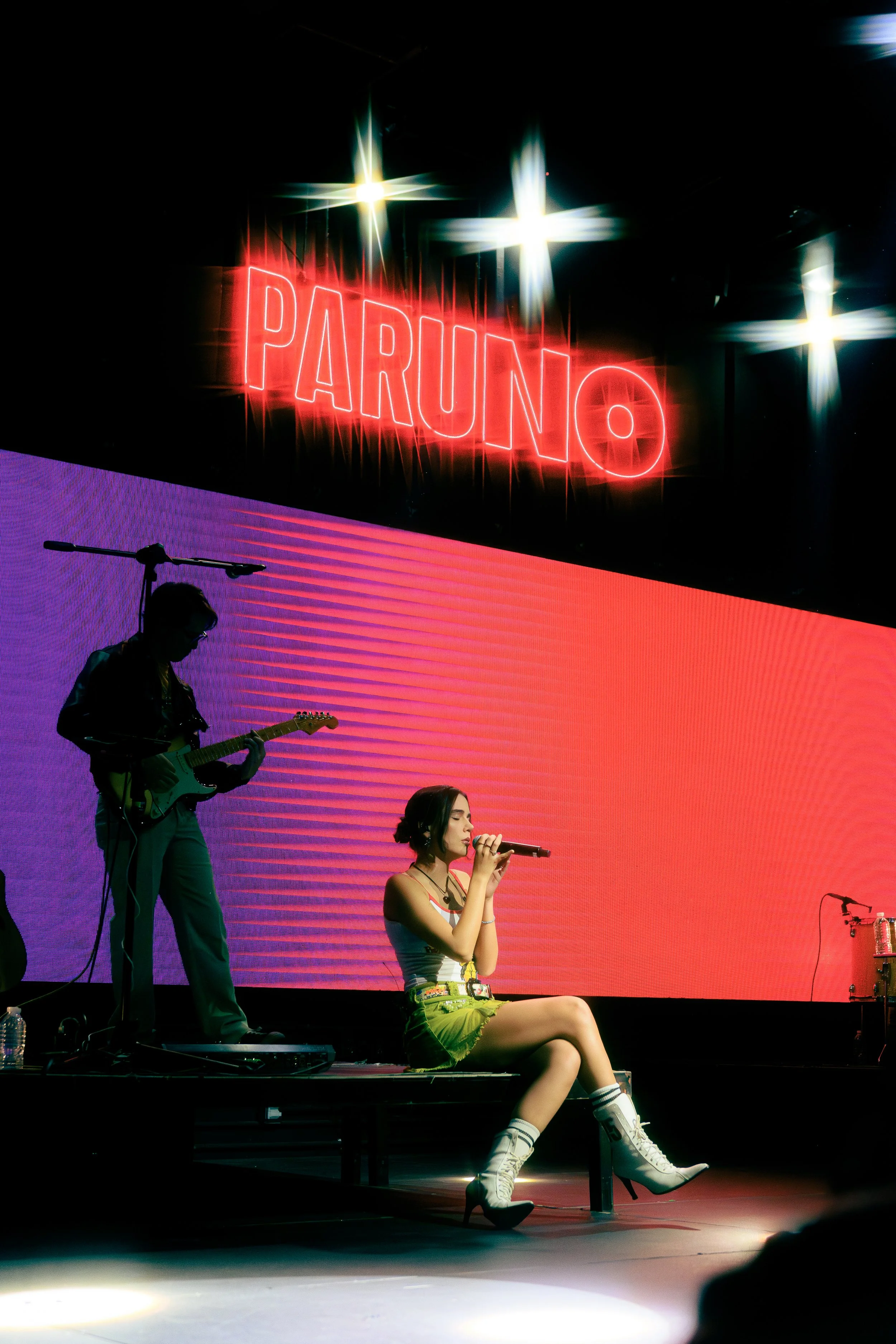 una cantante sentada en un escenario con un micrófono, acompañada por un músico tocando guitarra, con un letrero de neón rojo que dice 'PARUNO' y un fondo grande con colores degradados cercanos a tonos rosas y púrpuras.
